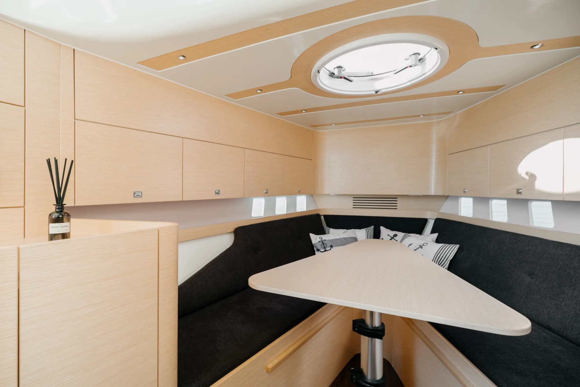The below-deck cabin of the Wia 330: a U-shaped charcoal dinette around a light oak table, pale oak panelling, a round overhead hatch and nautical anchor-print cushions.