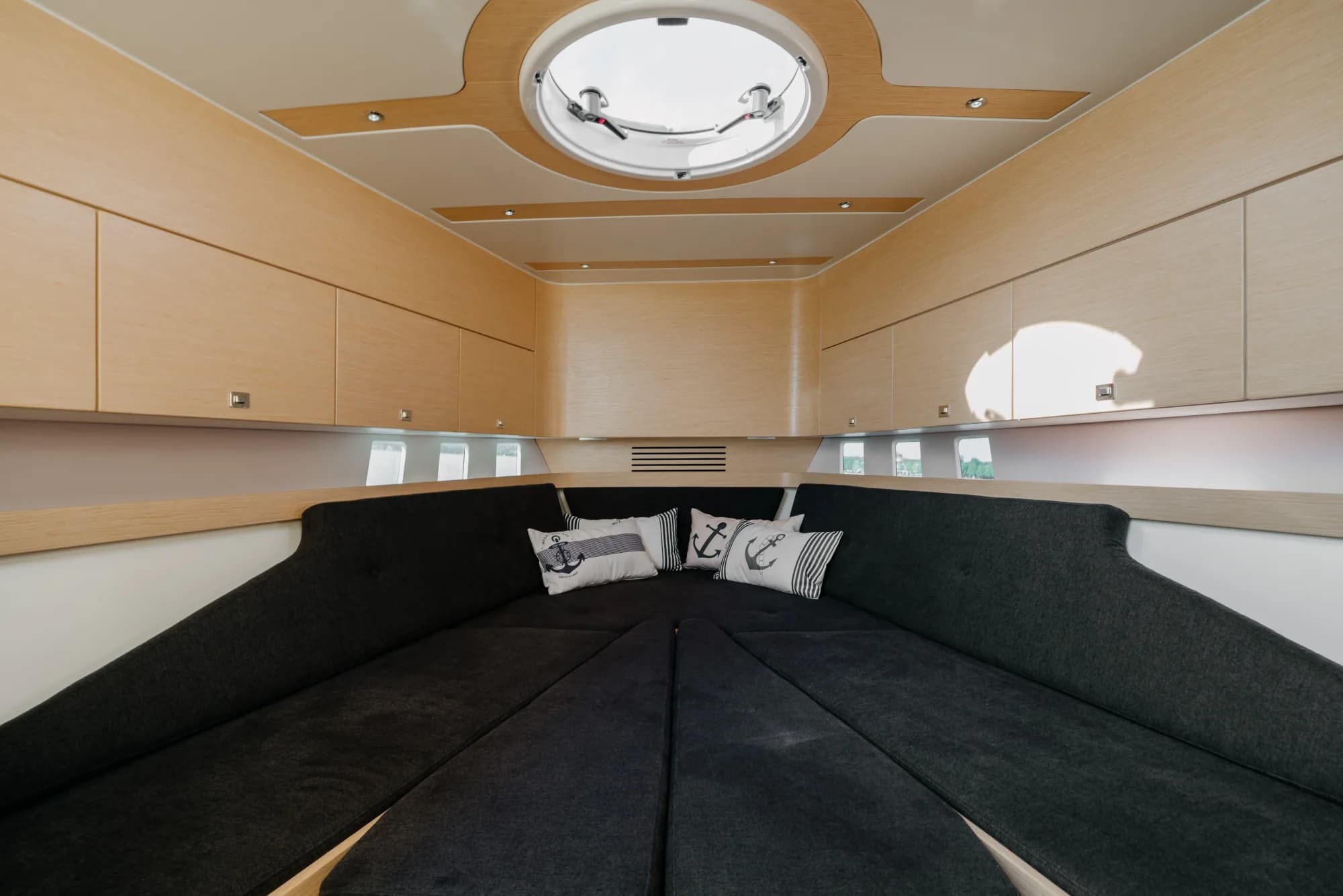 The below-deck cabin of the Wia 330: a U-shaped charcoal dinette around a light oak table, pale oak panelling, a round overhead hatch and nautical anchor-print cushions.