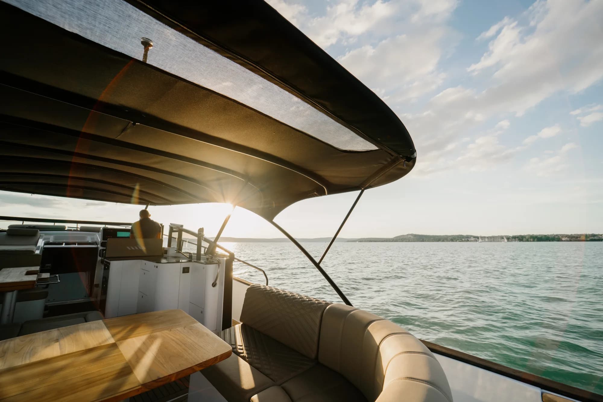The aft cockpit of the Wia 450 Cabrio at sunset: cream quilted-leather seating and a teak table under the black bimini top, a person at the helm and the hills of Lake Balaton beyond.