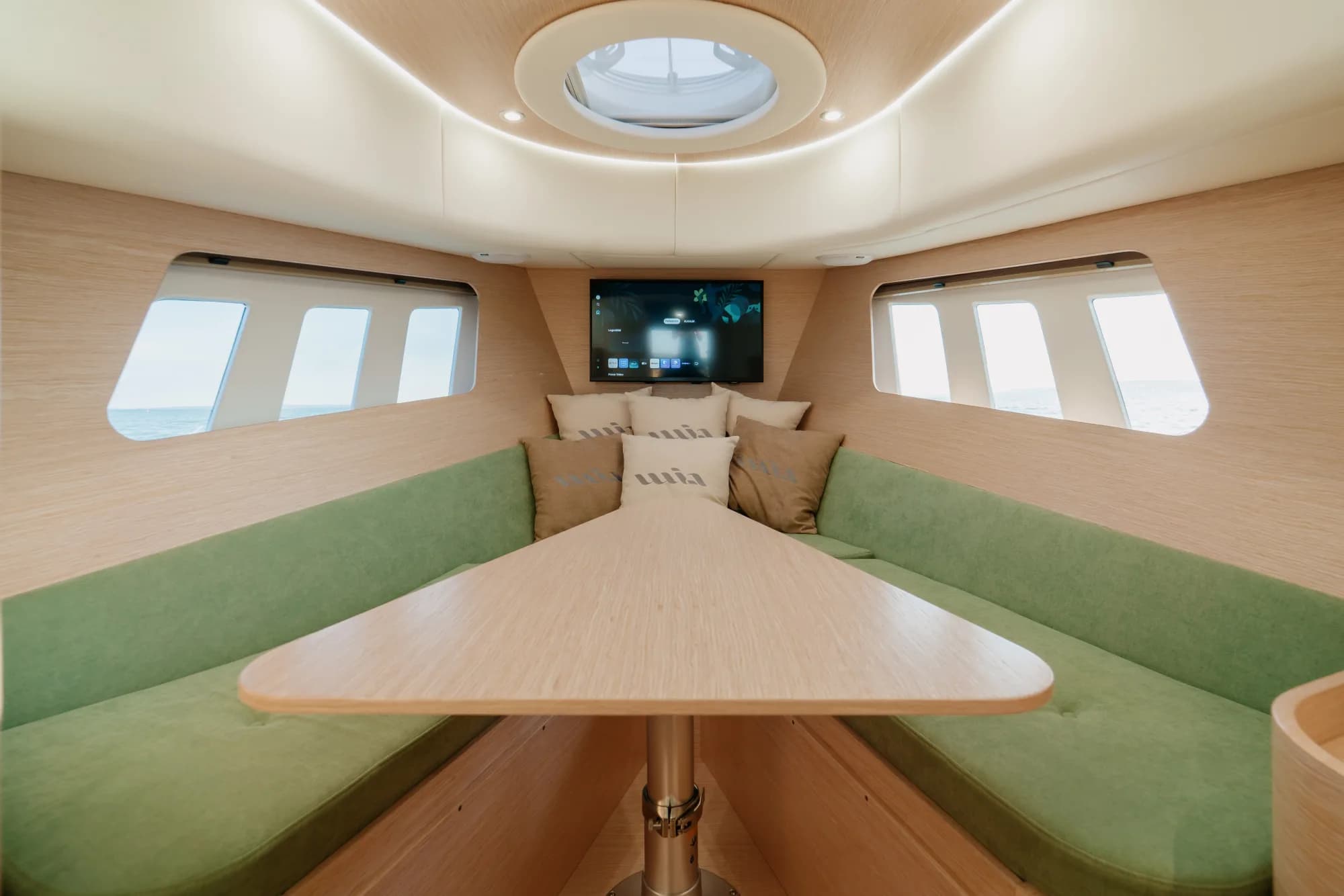 The saloon of the Wia 450 Cabrio: a U-shaped green dinette around a light oak table, pale oak panelling, a round overhead skylight, a wall-mounted screen and wia-branded cushions.