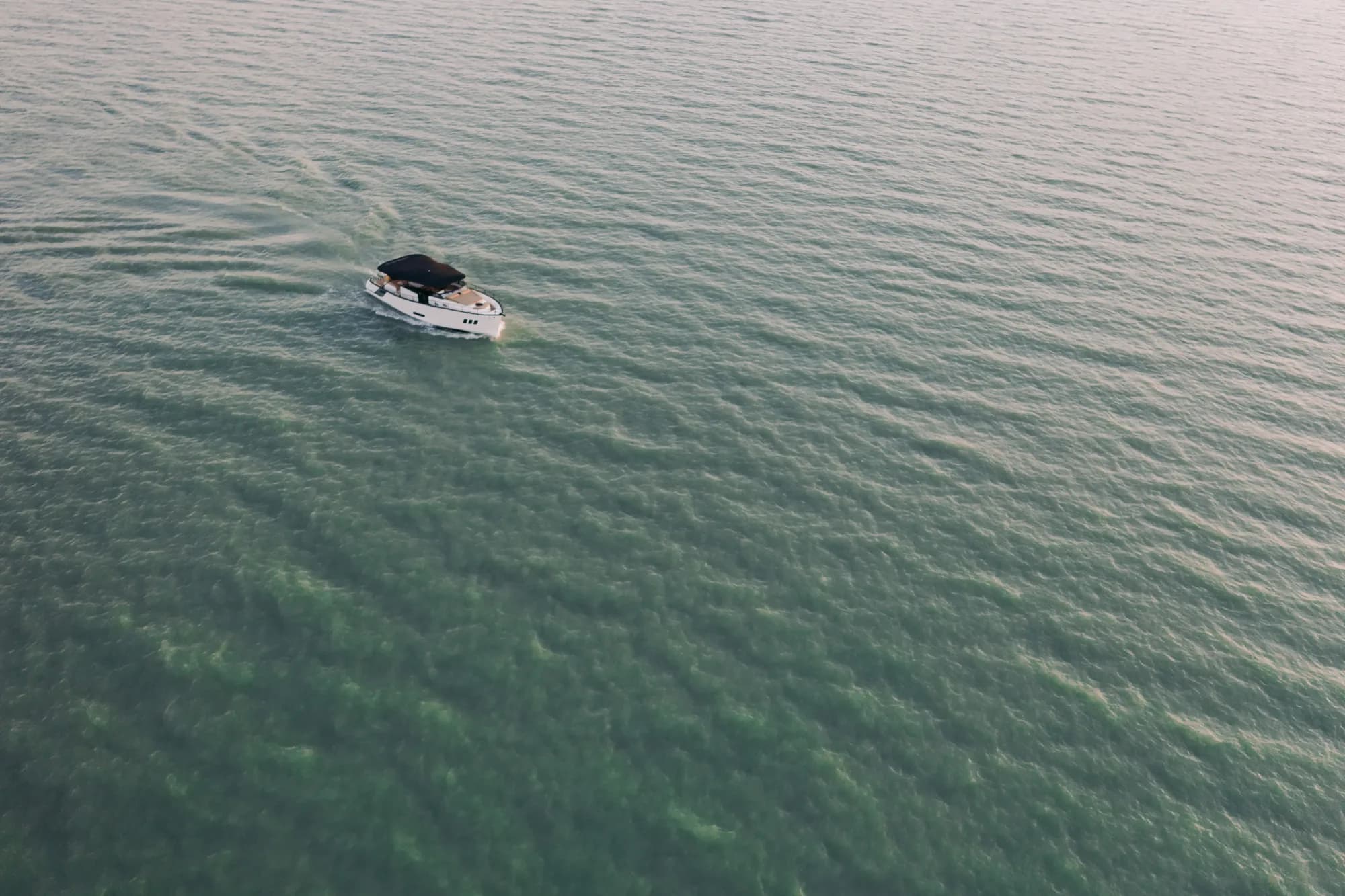 The Wia 450 Cabrio far below on shimmering green water, a small white boat with a dark canvas top leaving a faint wake.