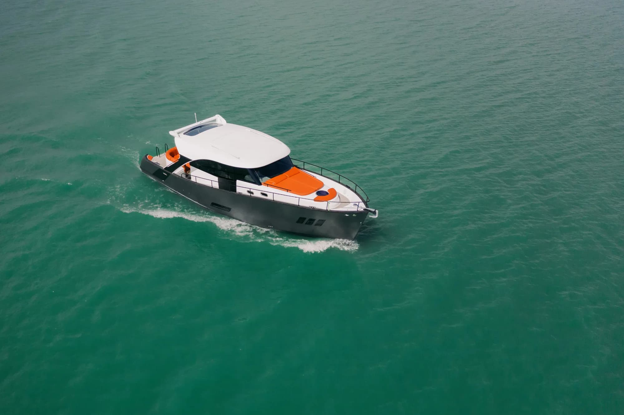 The dark-grey-hulled Wia 450 HardTop with its white hardtop and orange foredeck sunpads under way on turquoise water, seen from the air.