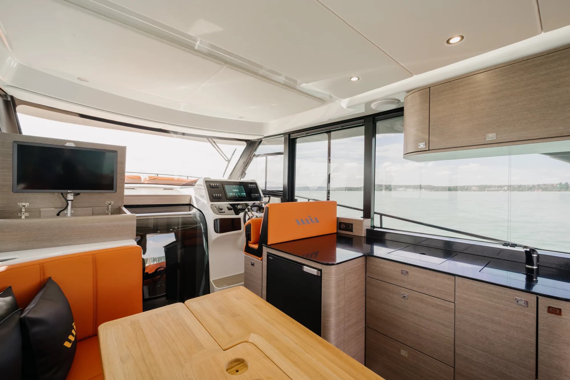 The enclosed saloon of the Wia 450 HardTop: orange leather seating with wia cushions, a light oak dinette, a galley with a dark worktop and the helm ahead, wrapped in panoramic windows.