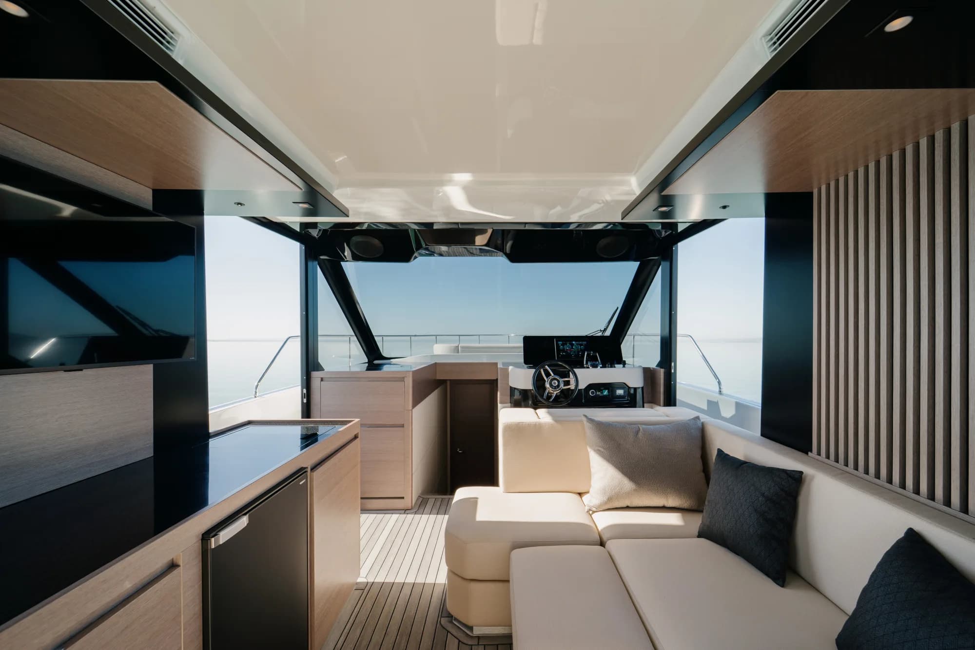 The enclosed saloon of the Wia 520 HardTop: a cream L-shaped sofa, light oak panelling, a galley with a dark worktop, a teak sole and the helm ahead, wrapped in panoramic windows.