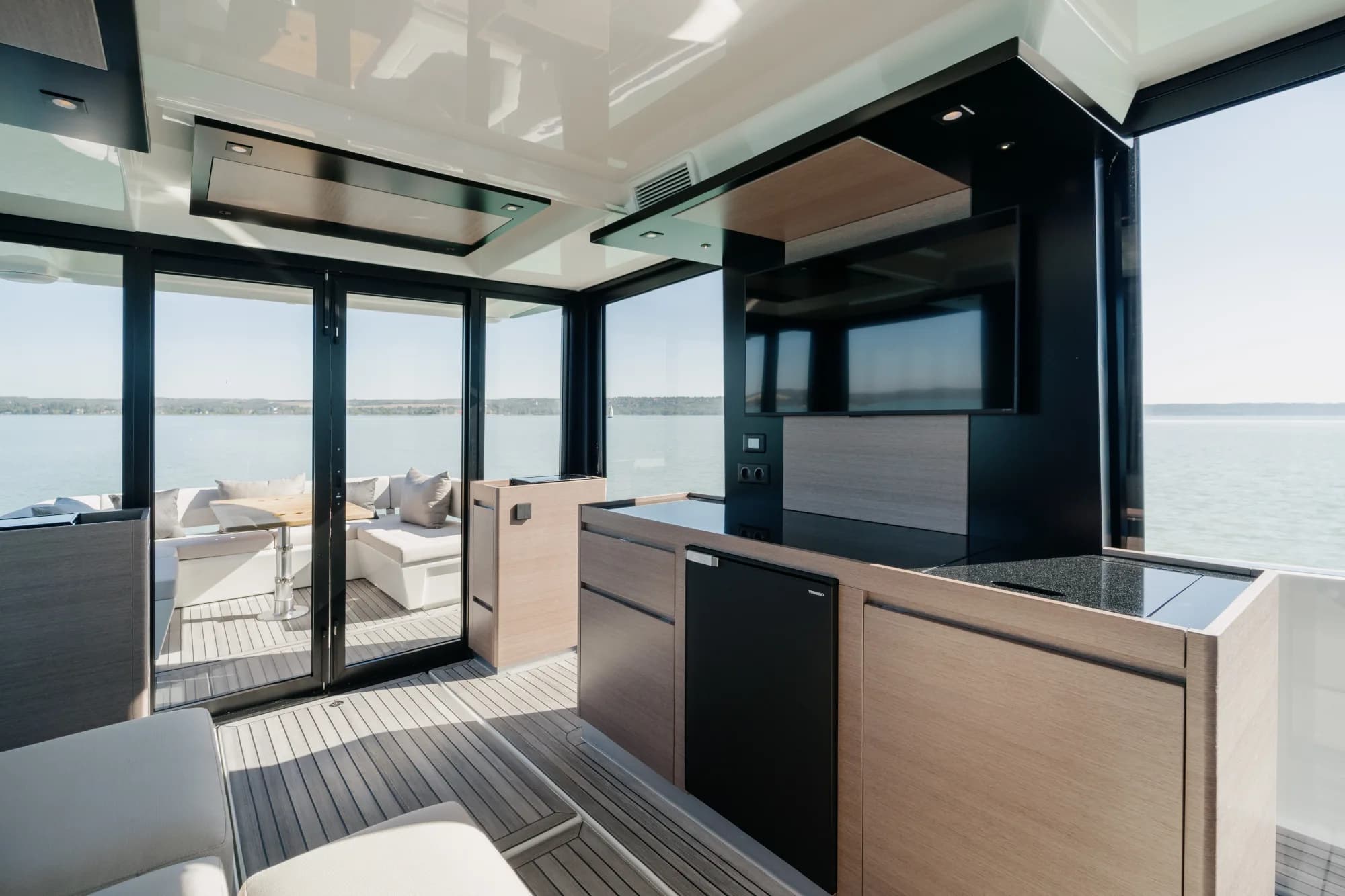 The enclosed saloon of the Wia 520 HardTop: a cream L-shaped sofa, light oak panelling, a galley with a dark worktop, a teak sole and the helm ahead, wrapped in panoramic windows.