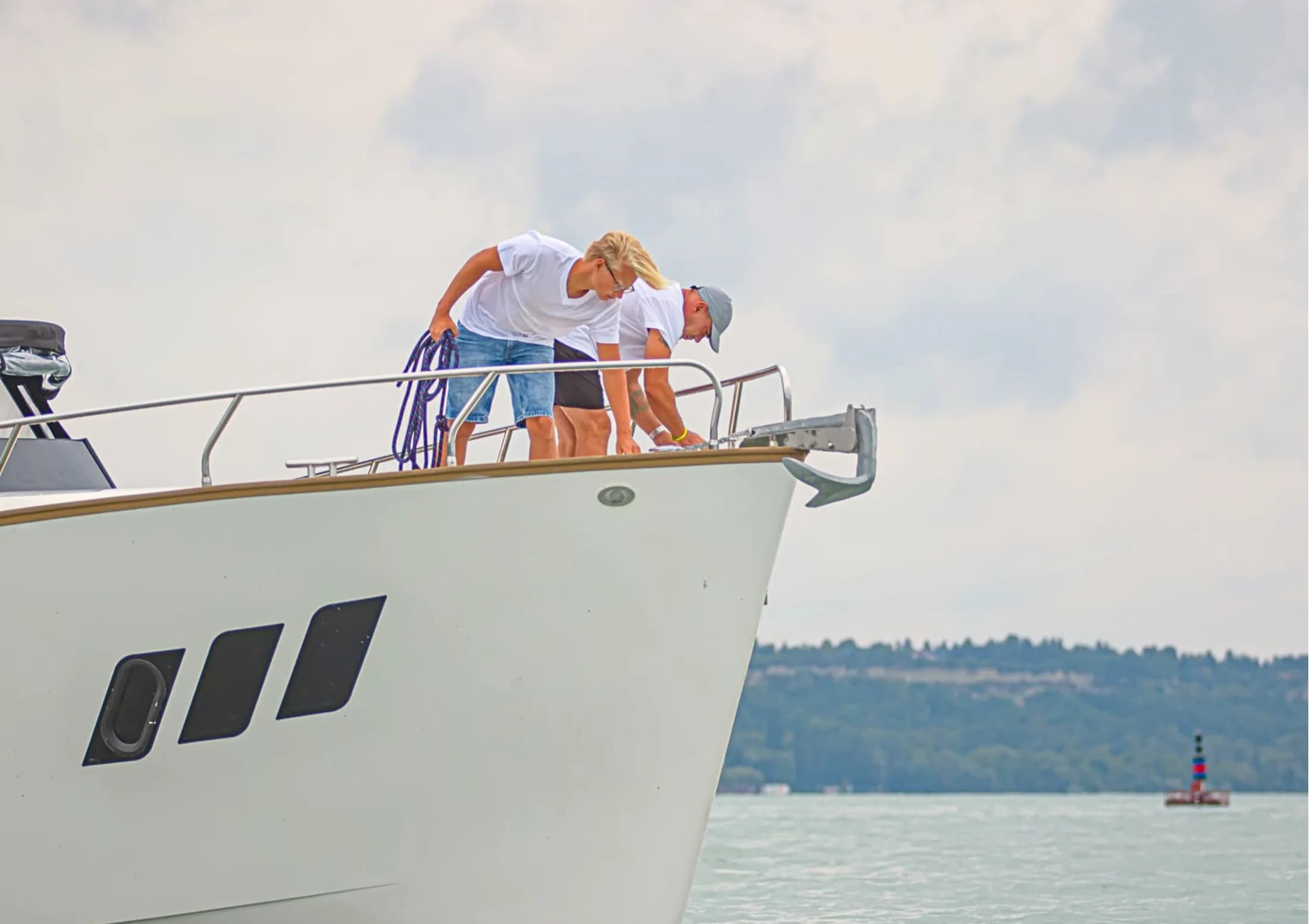 Two people on the bow of a white Wia yacht on the water, one leaning over the bow rail to handle a mooring line.