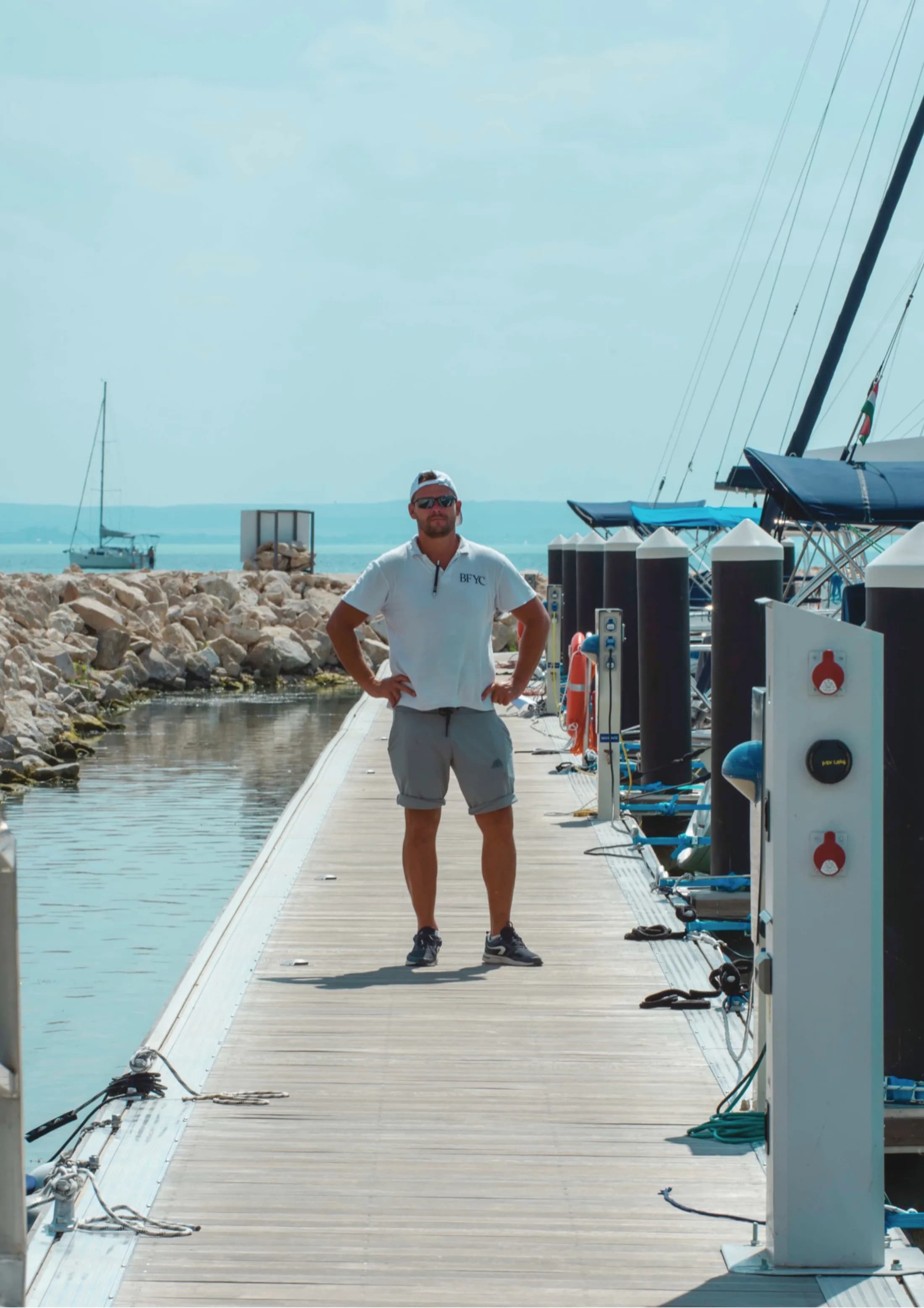 A man stands on a marina pontoon lined with shore-power pedestals, sailboats and the lake behind.