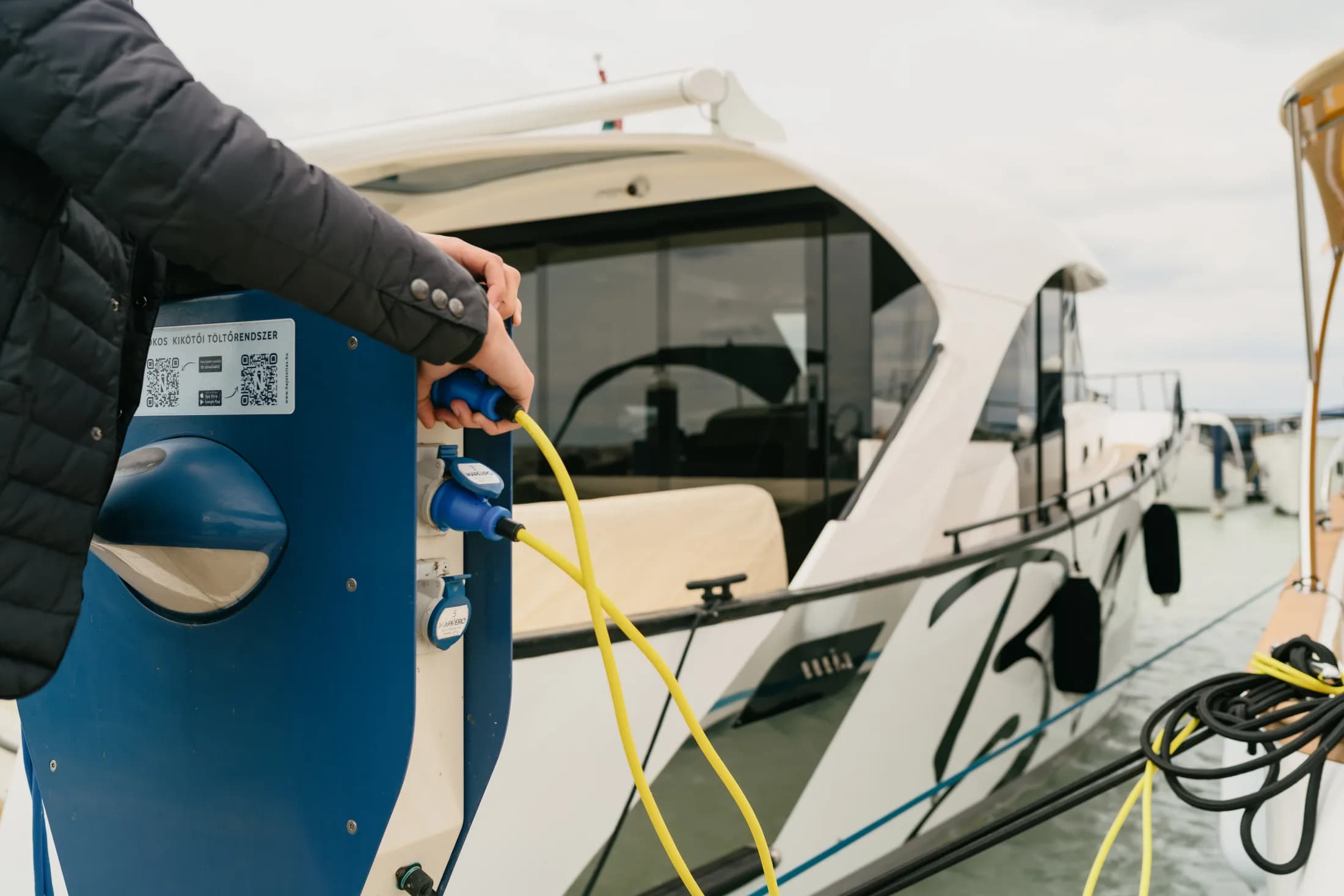 A person plugs a yellow shore-power cable into a blue marina charging pedestal beside a moored white Wia.