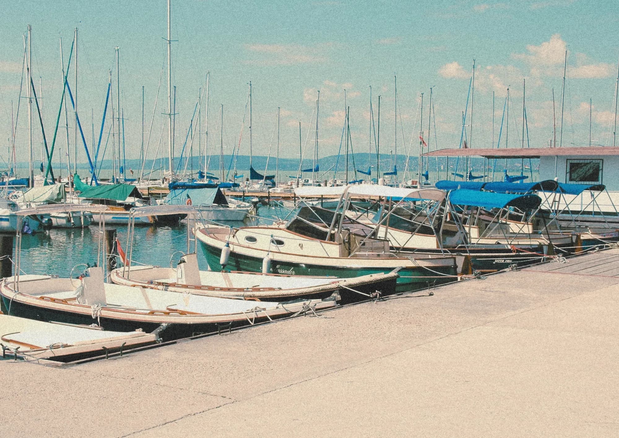 A Balaton marina lined with classic Wia launches and sailboats at their moorings, masts and hills beyond, in a warm vintage tone.