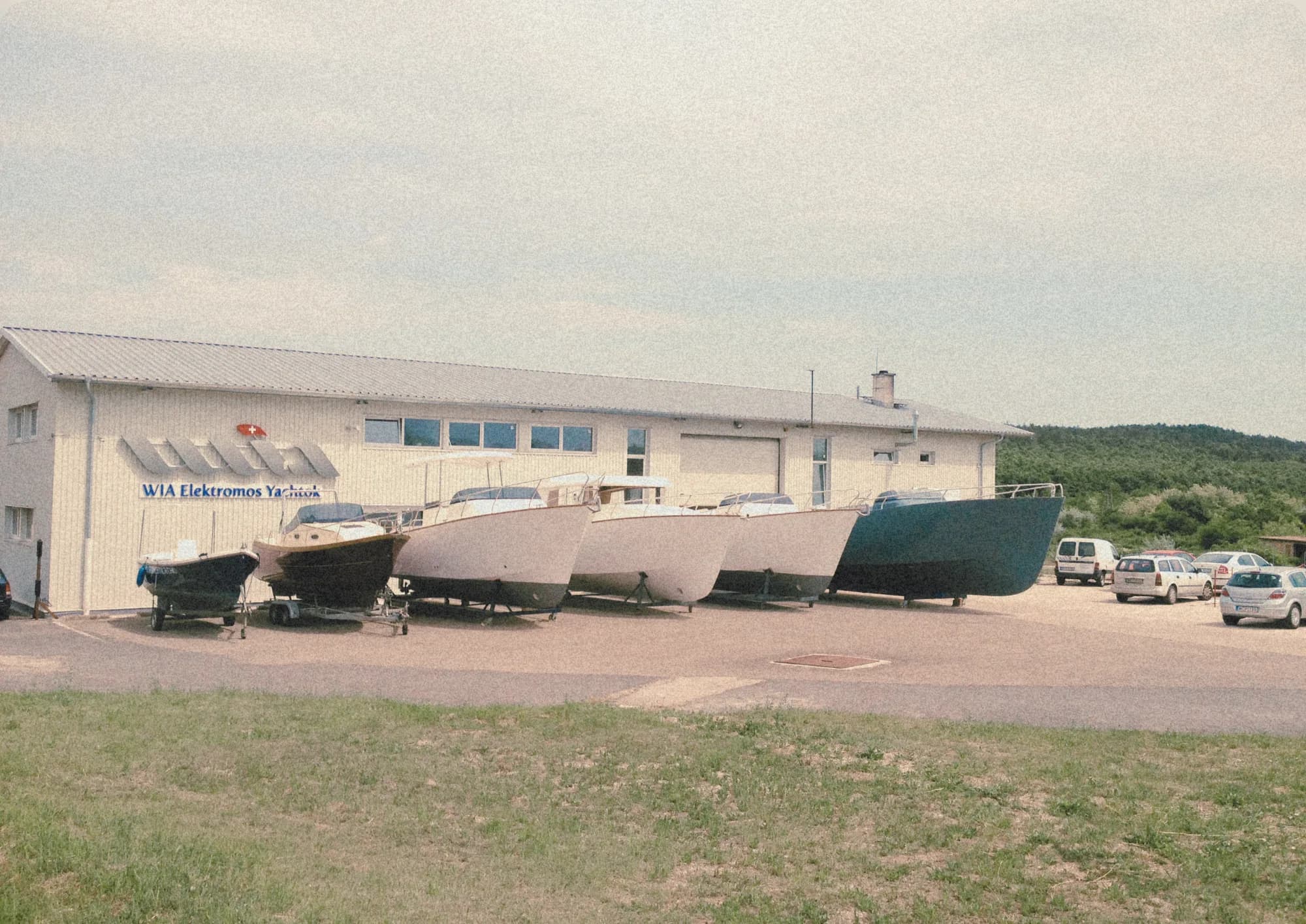 The 'WIA Elektromos Yachtok' workshop building with a row of hulls parked outside on trailers, wooded hills behind.