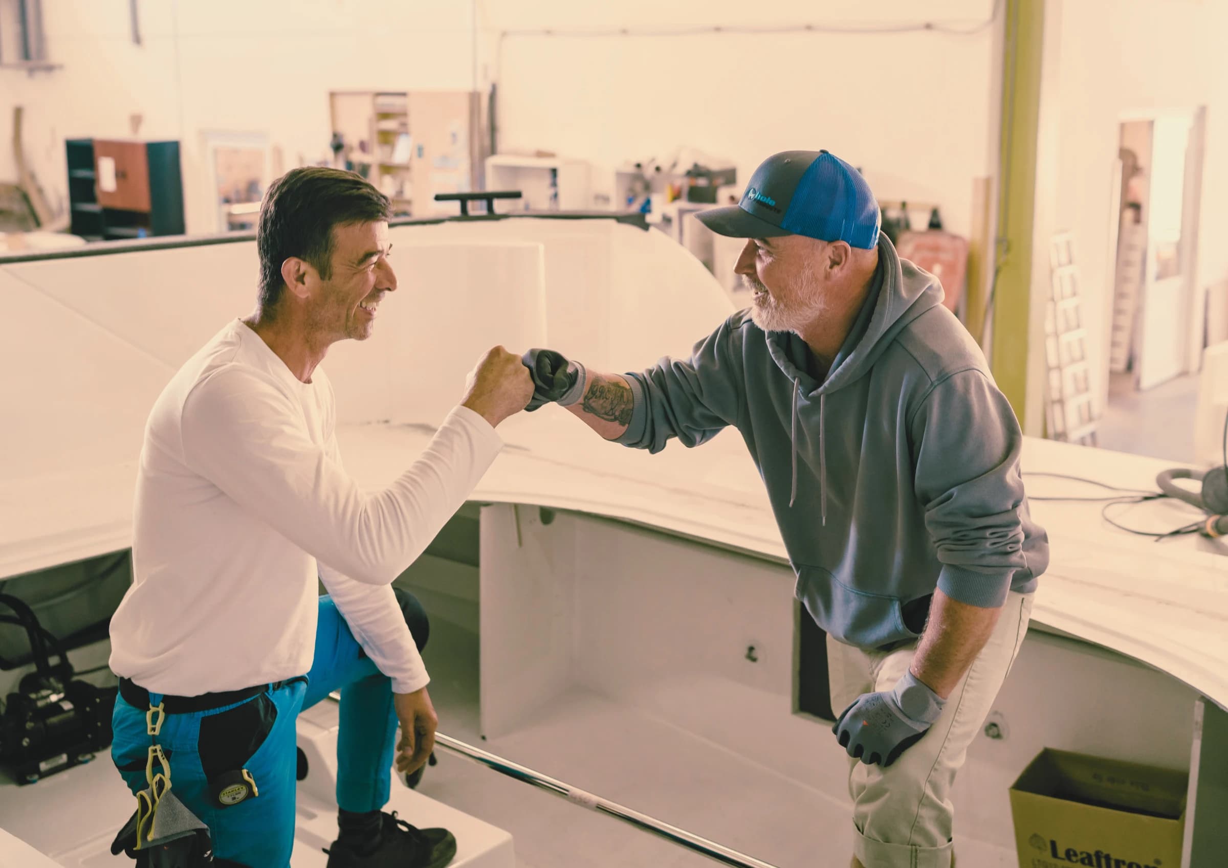 Two Wia boatbuilders bumping fists over a freshly laminated hull in the workshop.