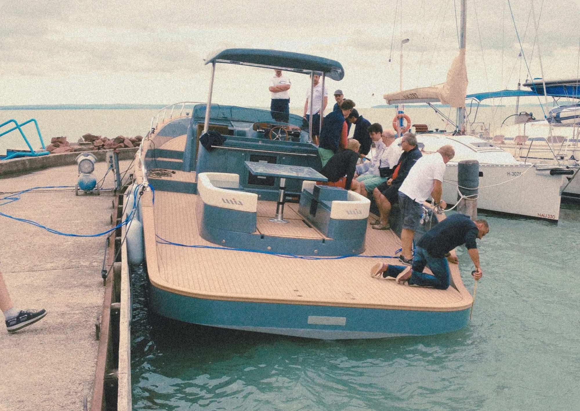 A blue Wia at a Balaton dock with a group aboard, one person kneeling to secure a line, in a warm vintage tone.