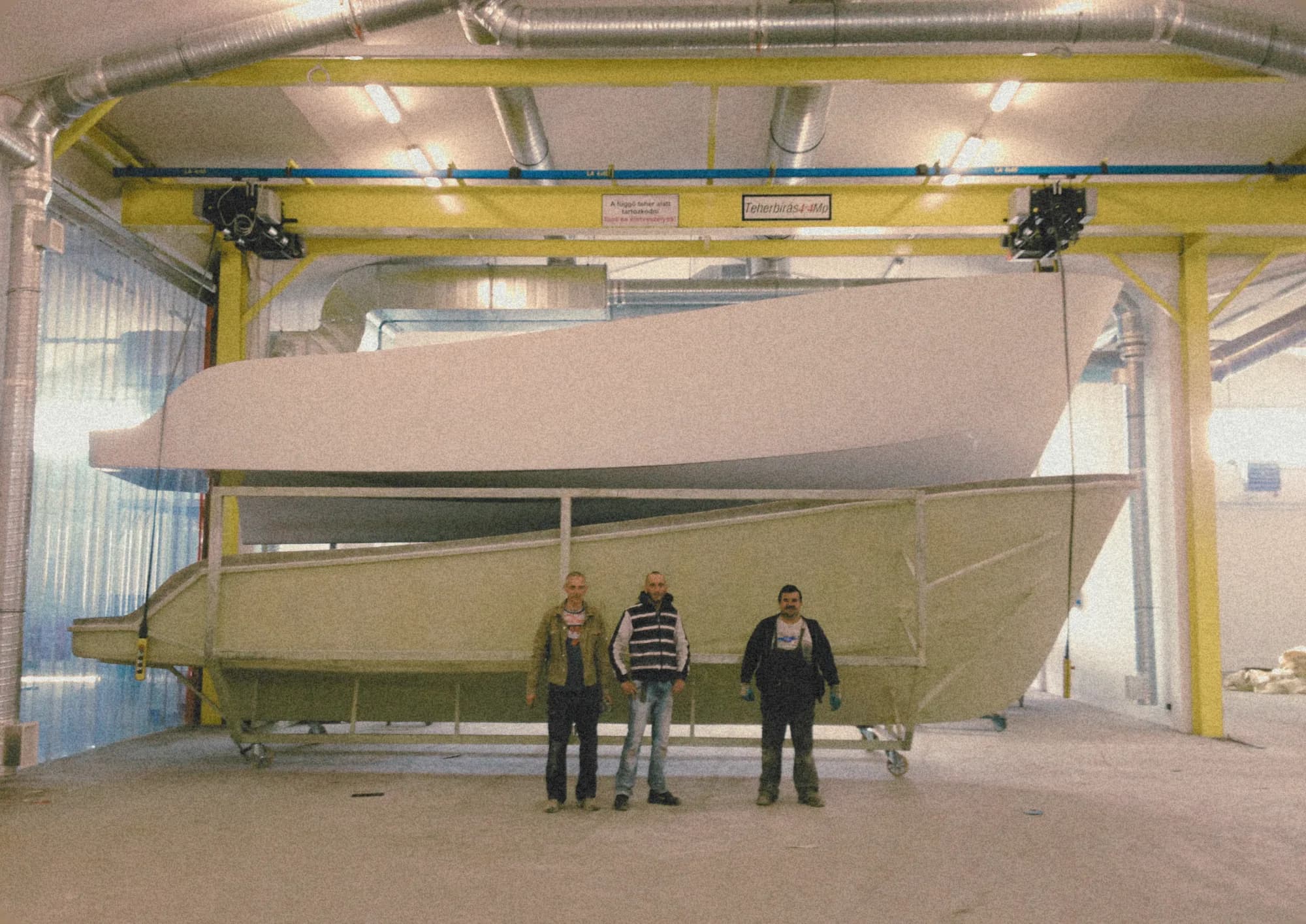 Three workers stand in front of a large Wia hull under construction inside the workshop, an overhead crane above.