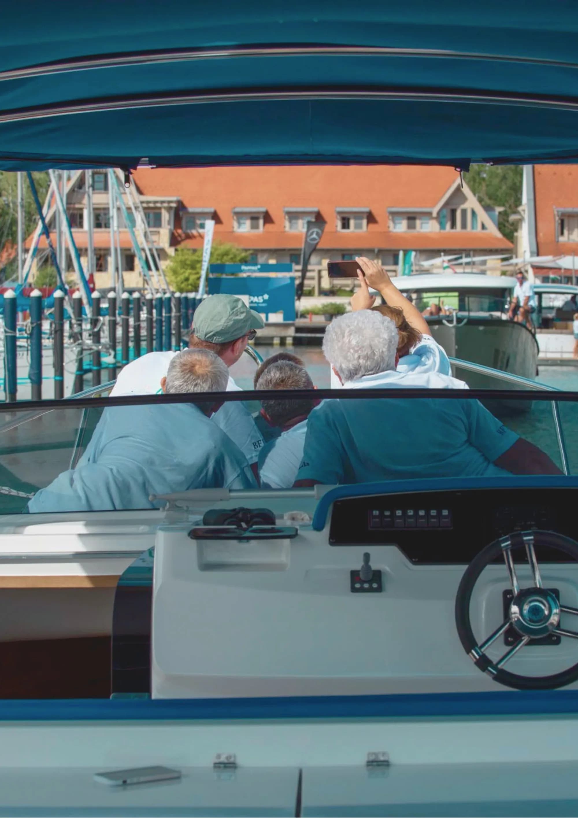 View forward from a Wia's cockpit under a blue bimini, a group of guests aboard and one taking a photo, marina buildings behind.