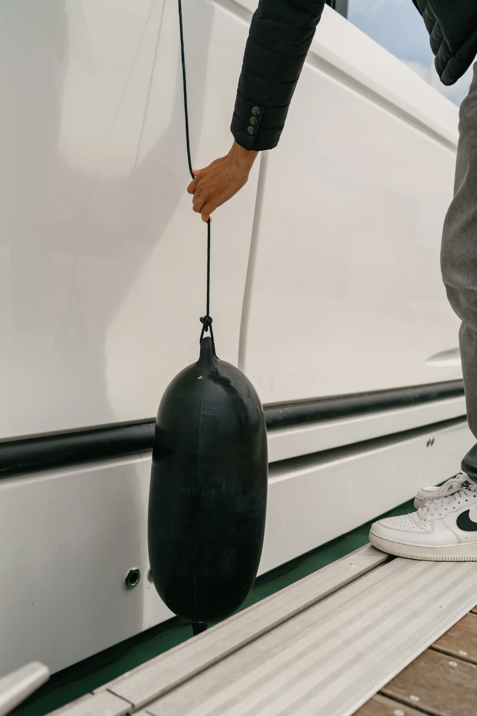 A person lowers a black fender on a line over the white hull of a Wia moored at a wooden dock.