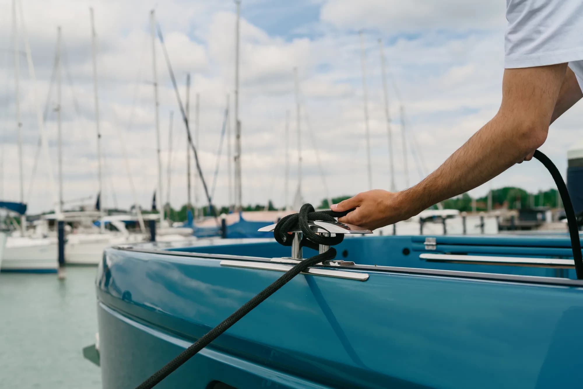 A hand securing a black mooring line on the stainless cleat of a teal-hulled Wia, sailboat masts behind.