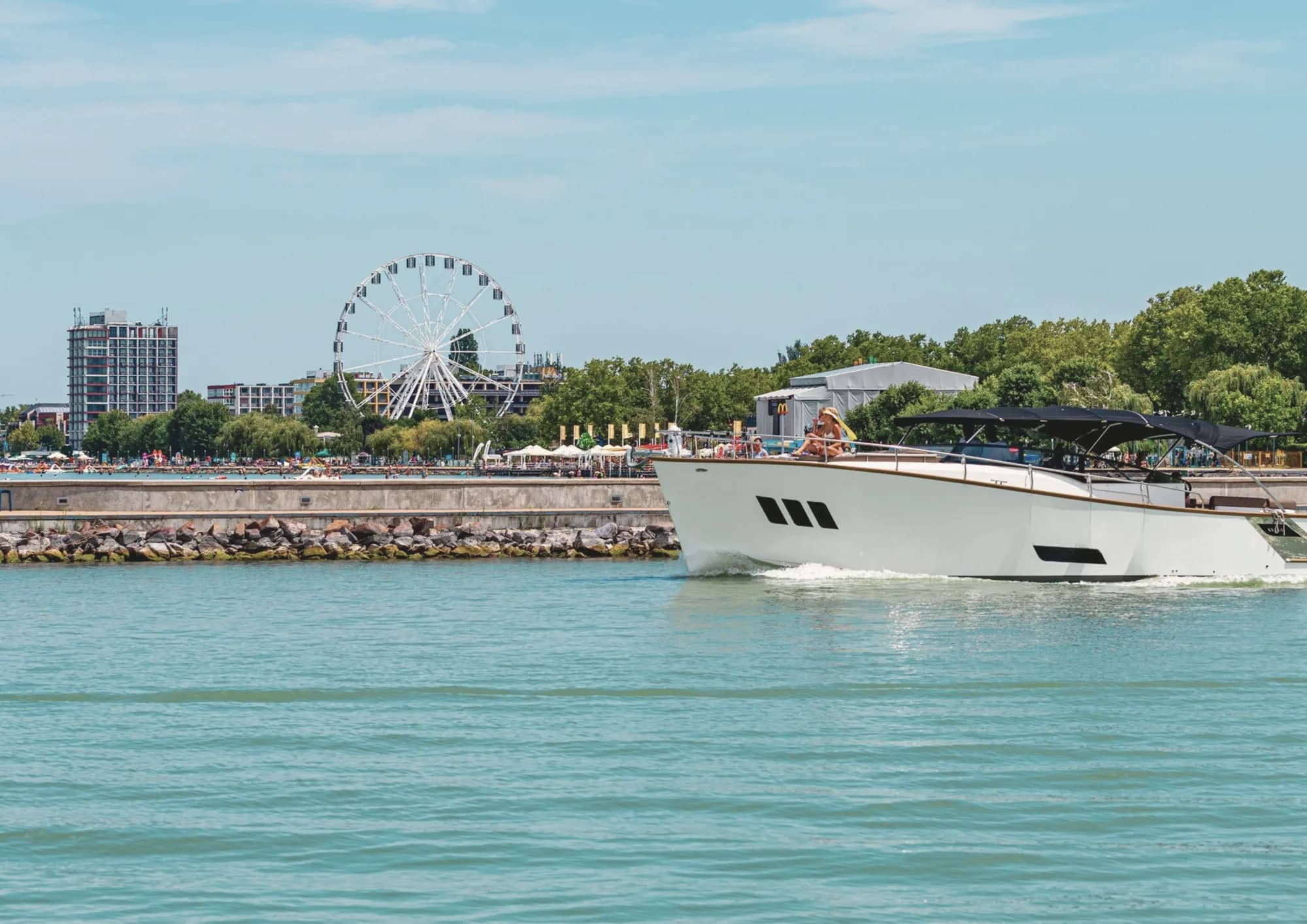 A white Wia cruises past a lakeside promenade with a Ferris wheel and a busy beach, a couple relaxing on the bow.