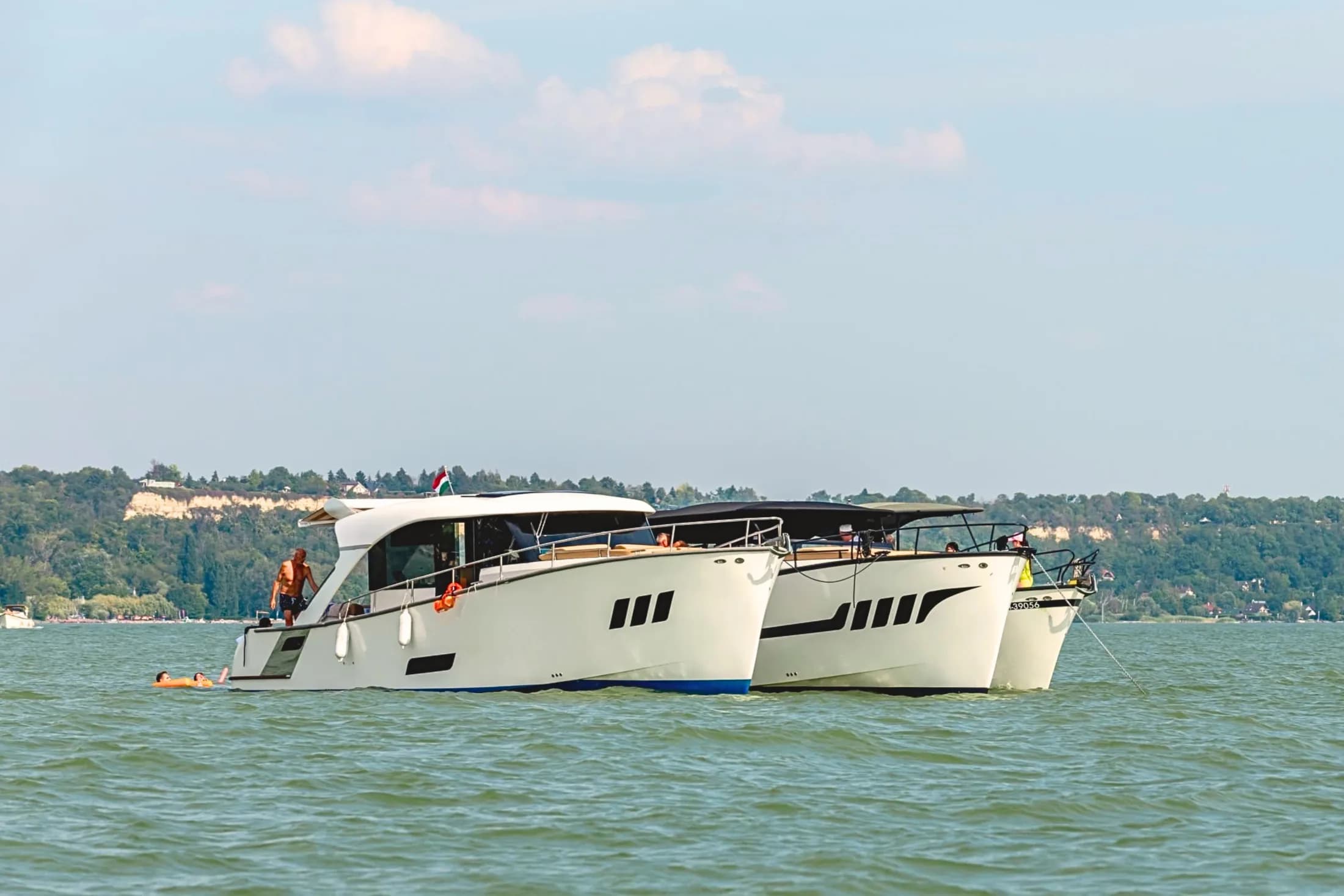 Two white Wia yachts side by side on Lake Balaton, a man on the side deck and two people on an orange float, Hungarian flag flying.