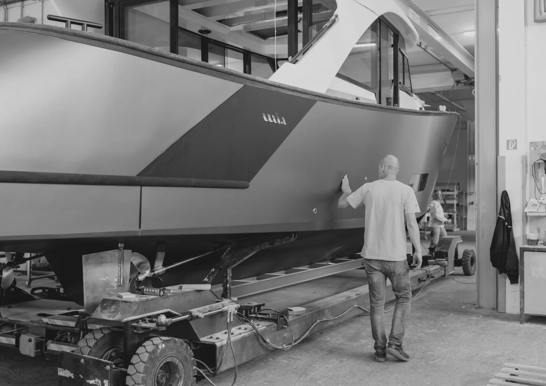 Photo en noir et blanc d'une coque Wia sur son ber dans l'atelier, un ouvrier marchant à ses côtés.