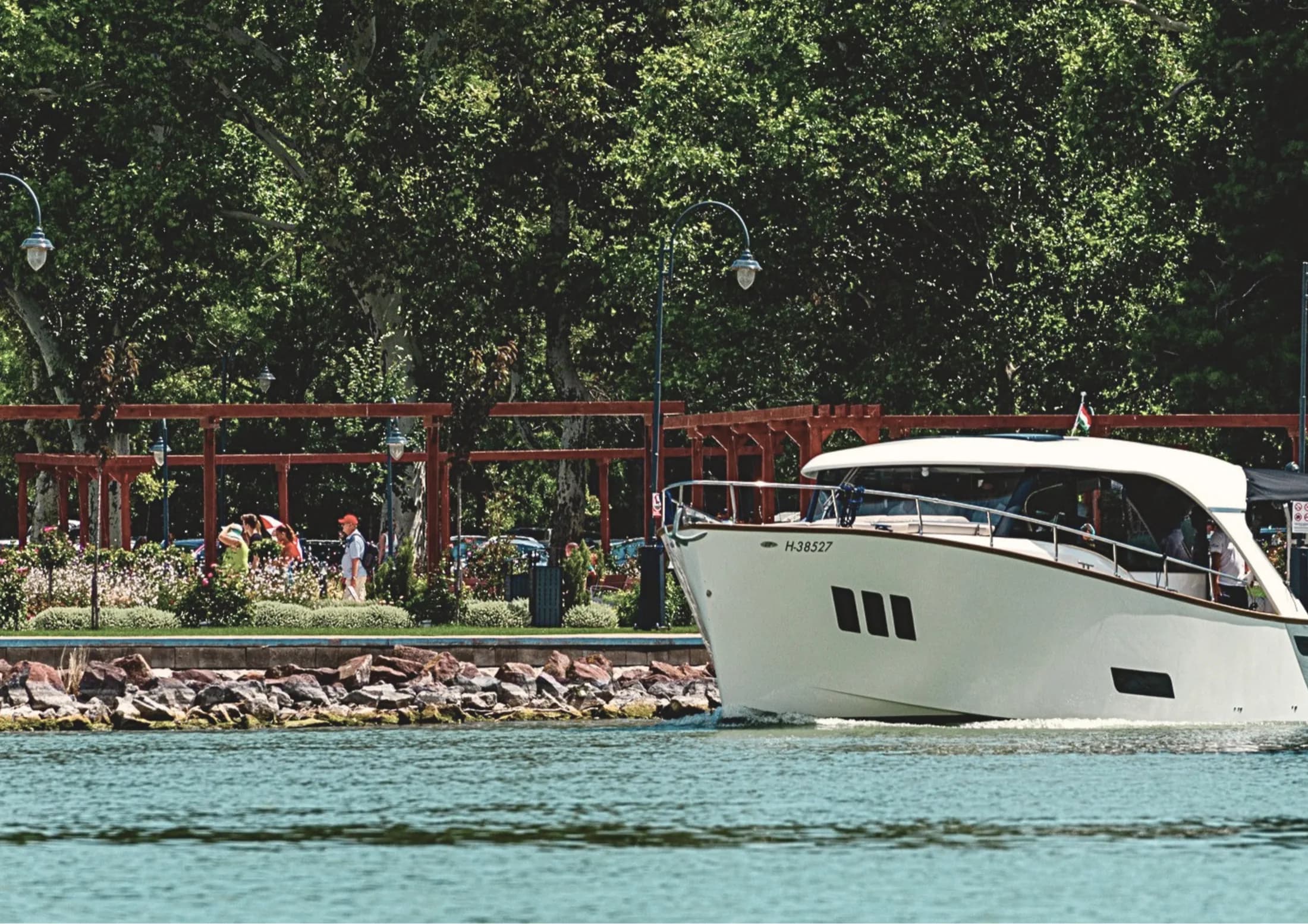 Un yacht Wia hardtop blanc navigue sur l’eau le long d’une promenade verdoyante au bord du lac, avec des pergolas rouges et des passants, un drapeau hongrois à la poupe.