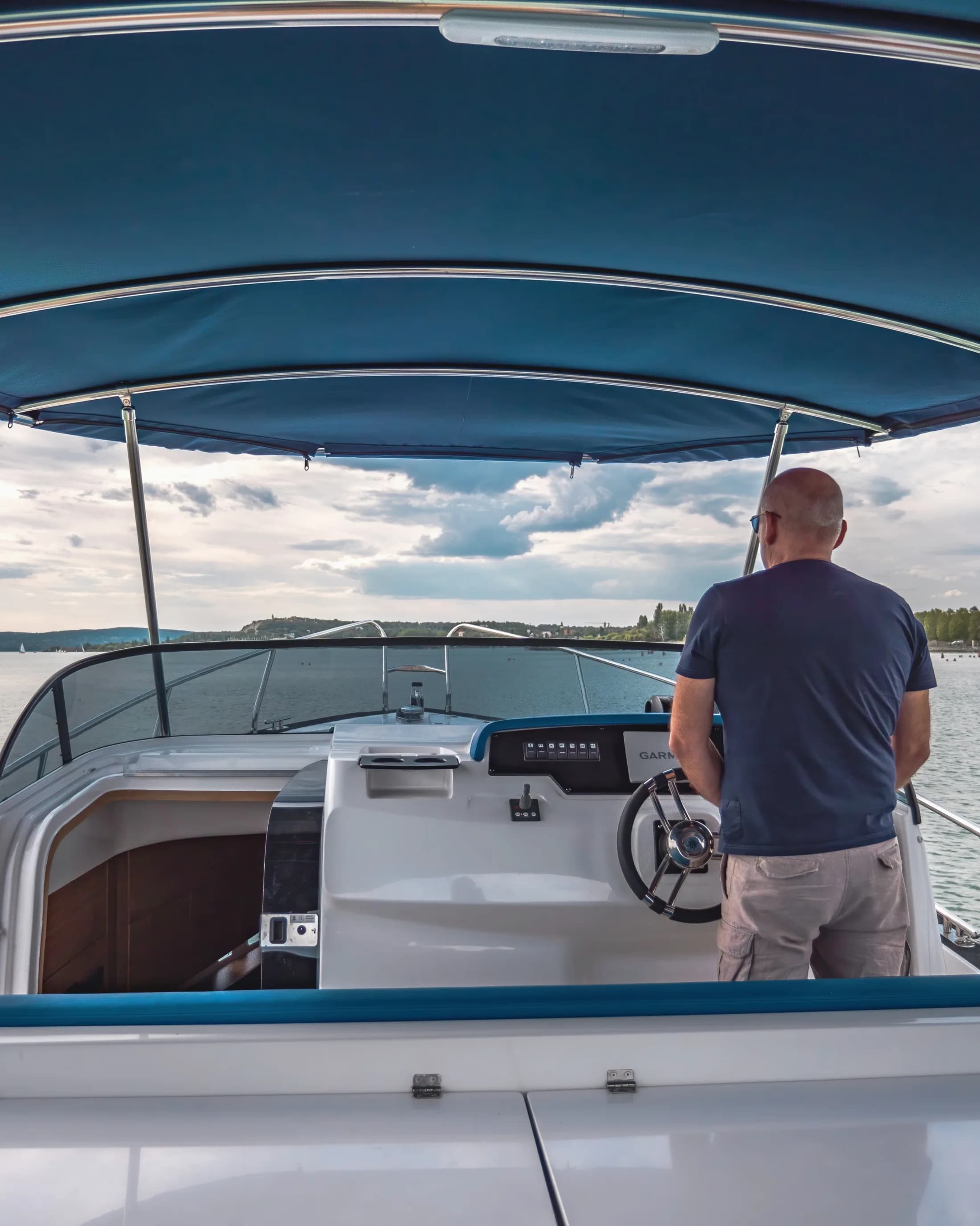 Un homme à la barre d'un Wia sous un bimini bleu, volant et écran Garmin, regardant le lac paisible.