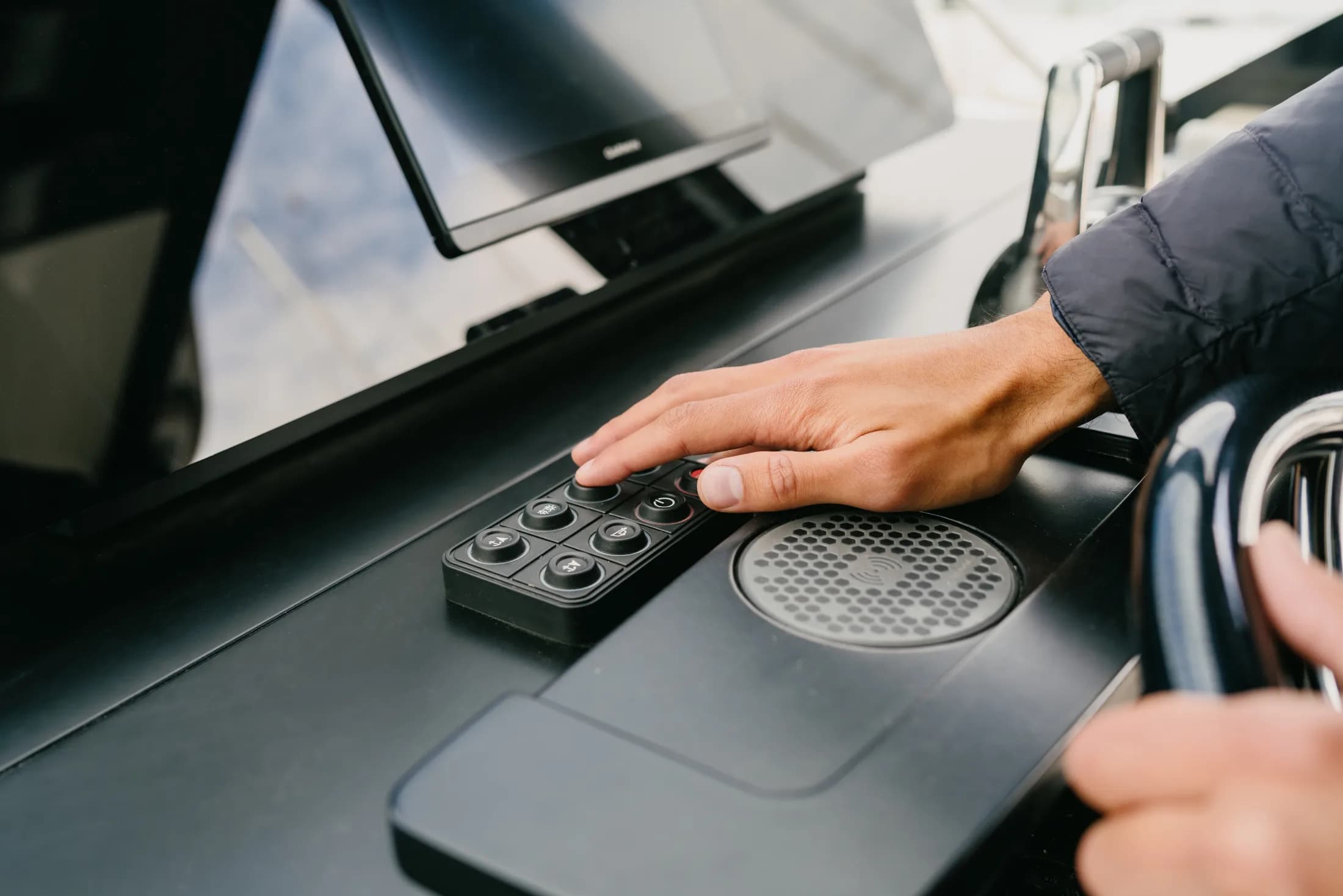 Une main sur un clavier de commande noir au poste de pilotage, près d'un écran Garmin et d'un chargeur sans fil.