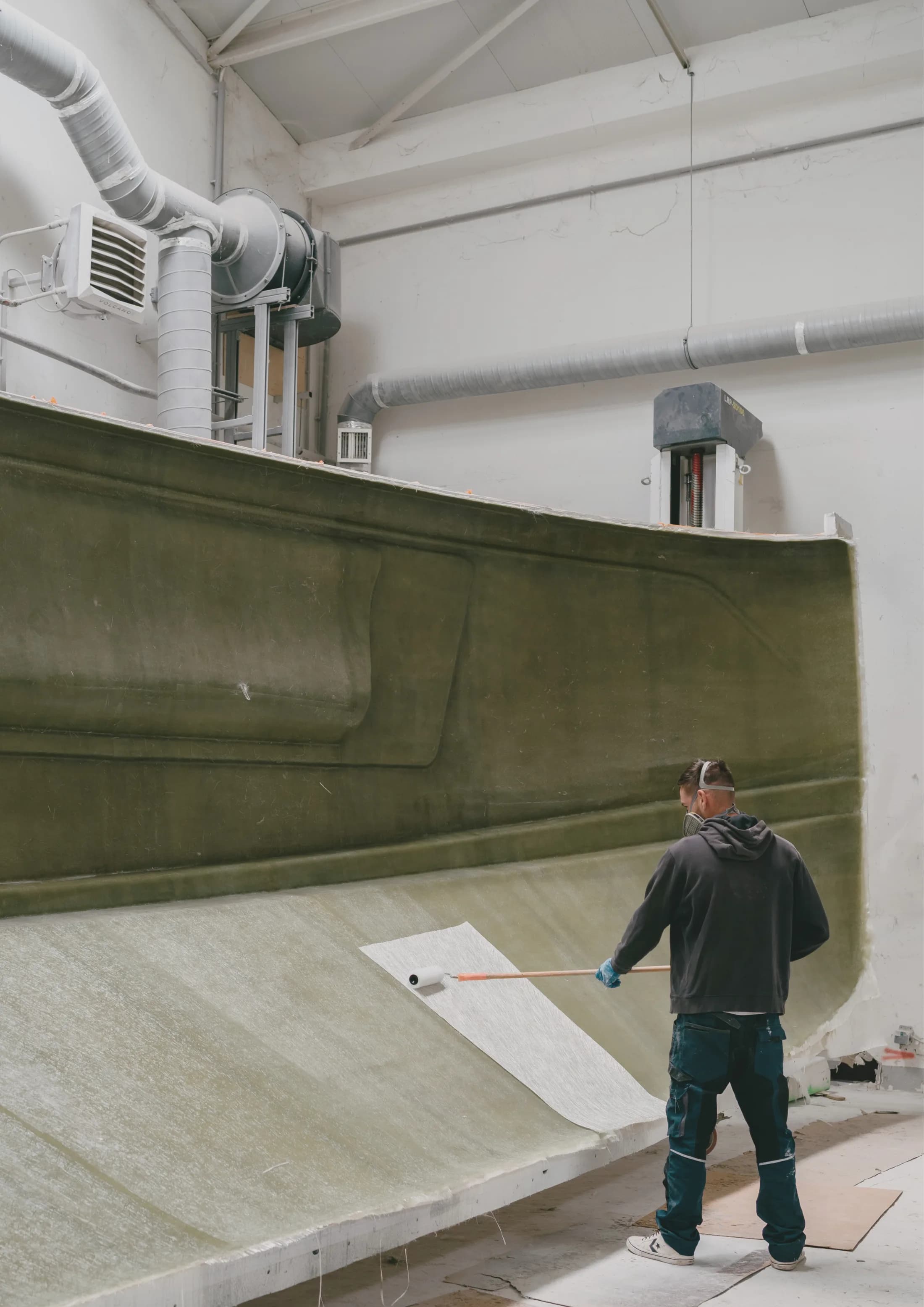 Un ouvrier muni d'un masque stratifie à la main de la fibre de verre sur un grand moule de coque vert dans l'atelier.