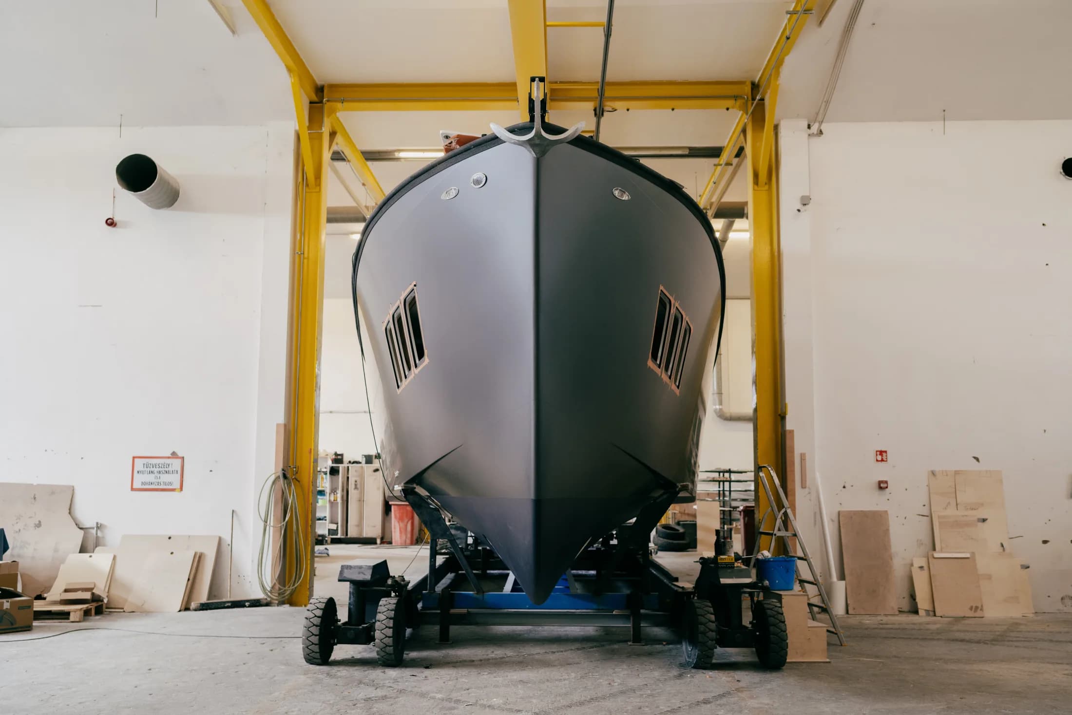 Un yacht Wia à coque grise vu de face dans l'atelier sur un ber à roulettes, sous un pont roulant jaune.