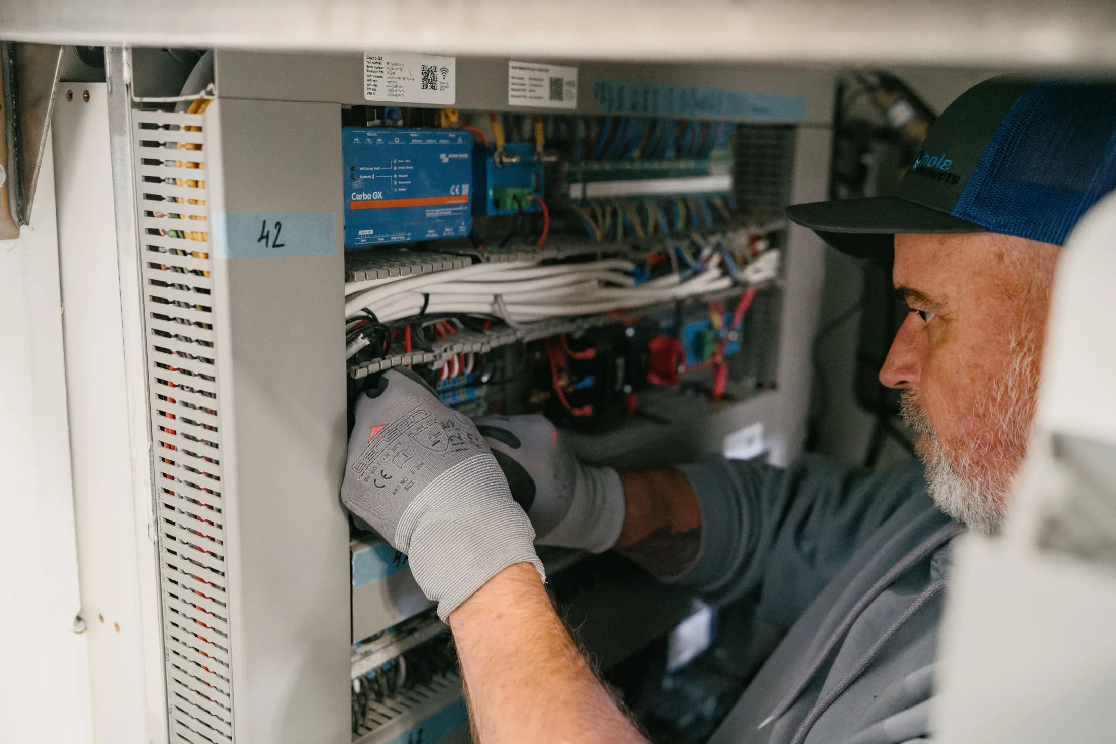 Un technicien travaille sur le système électrique du bateau, une main gantée sur le câblage derrière un Cerbo GX.