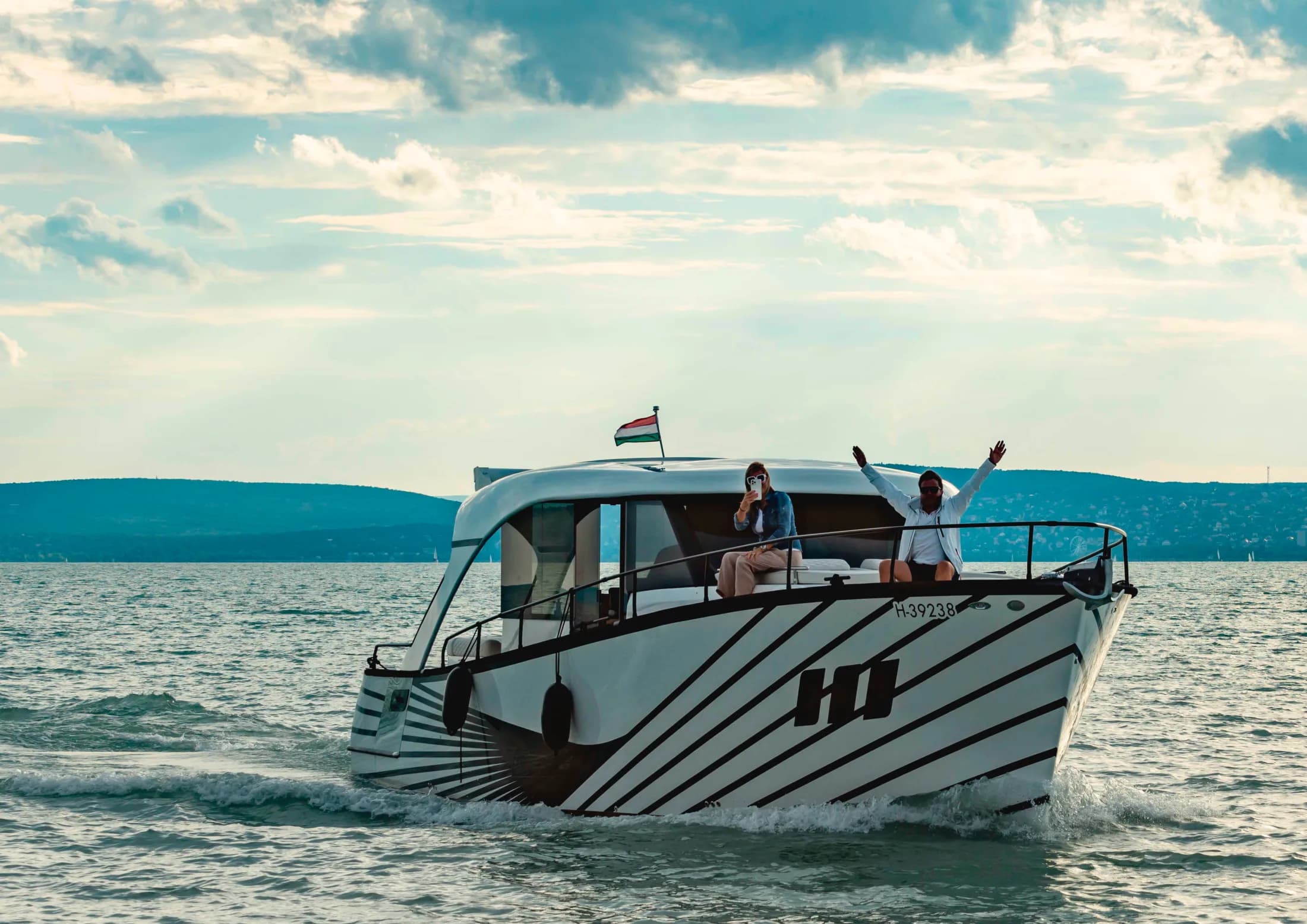 A white Wia with a striped 'HI' livery cruising on the lake, a woman photographing and a man with arms raised on the bow, Hungarian flag flying.