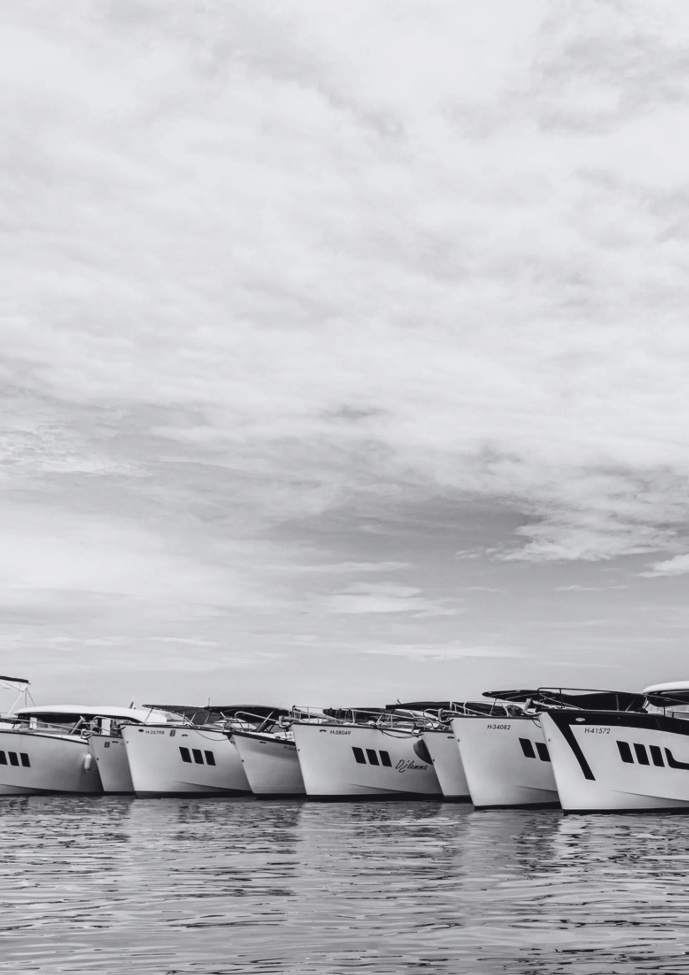 Black-and-white photo of a row of Wia yachts moored bow to bow on the water under a cloudy sky.