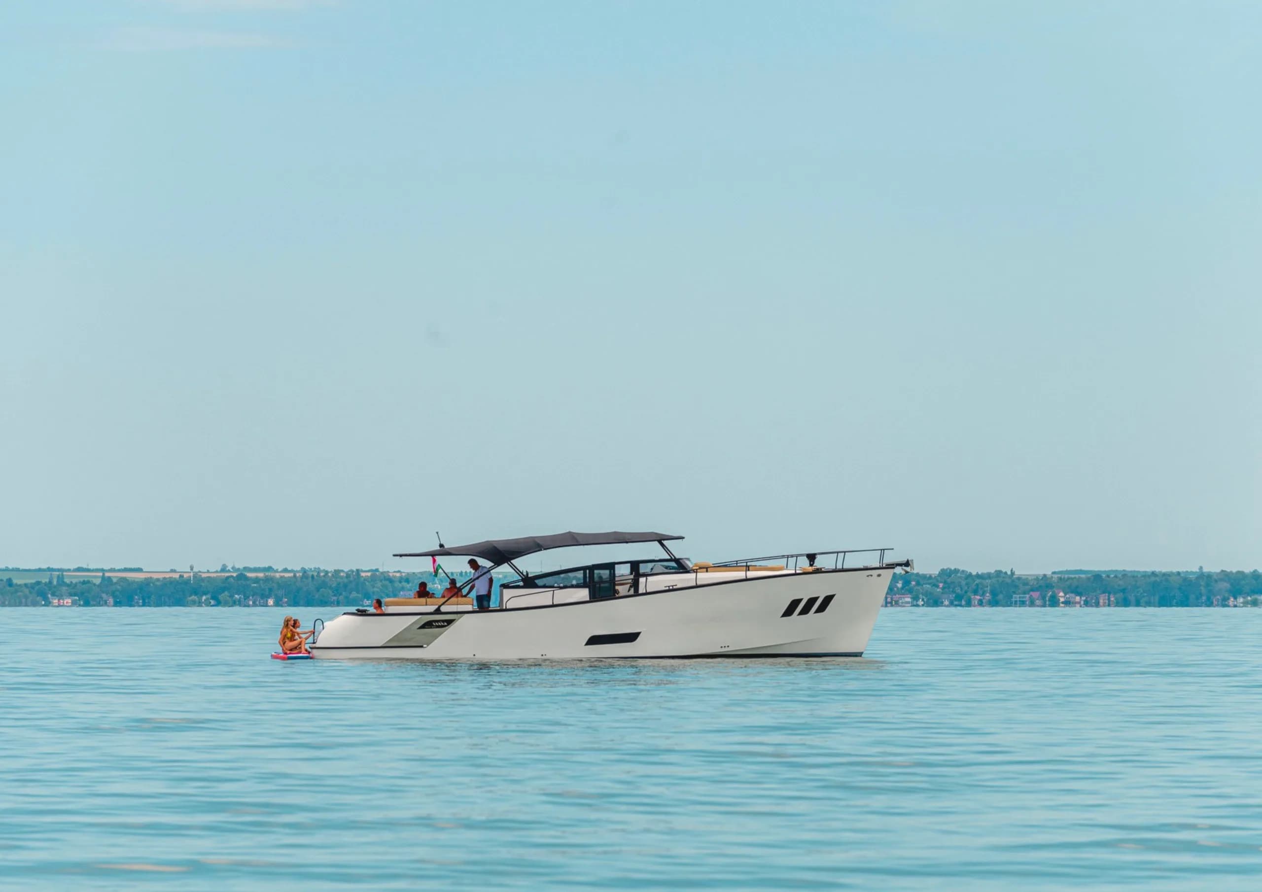 A white Wia yacht cruising in profile on a calm blue lake, guests under a dark bimini top and two people on a paddleboard at the stern.