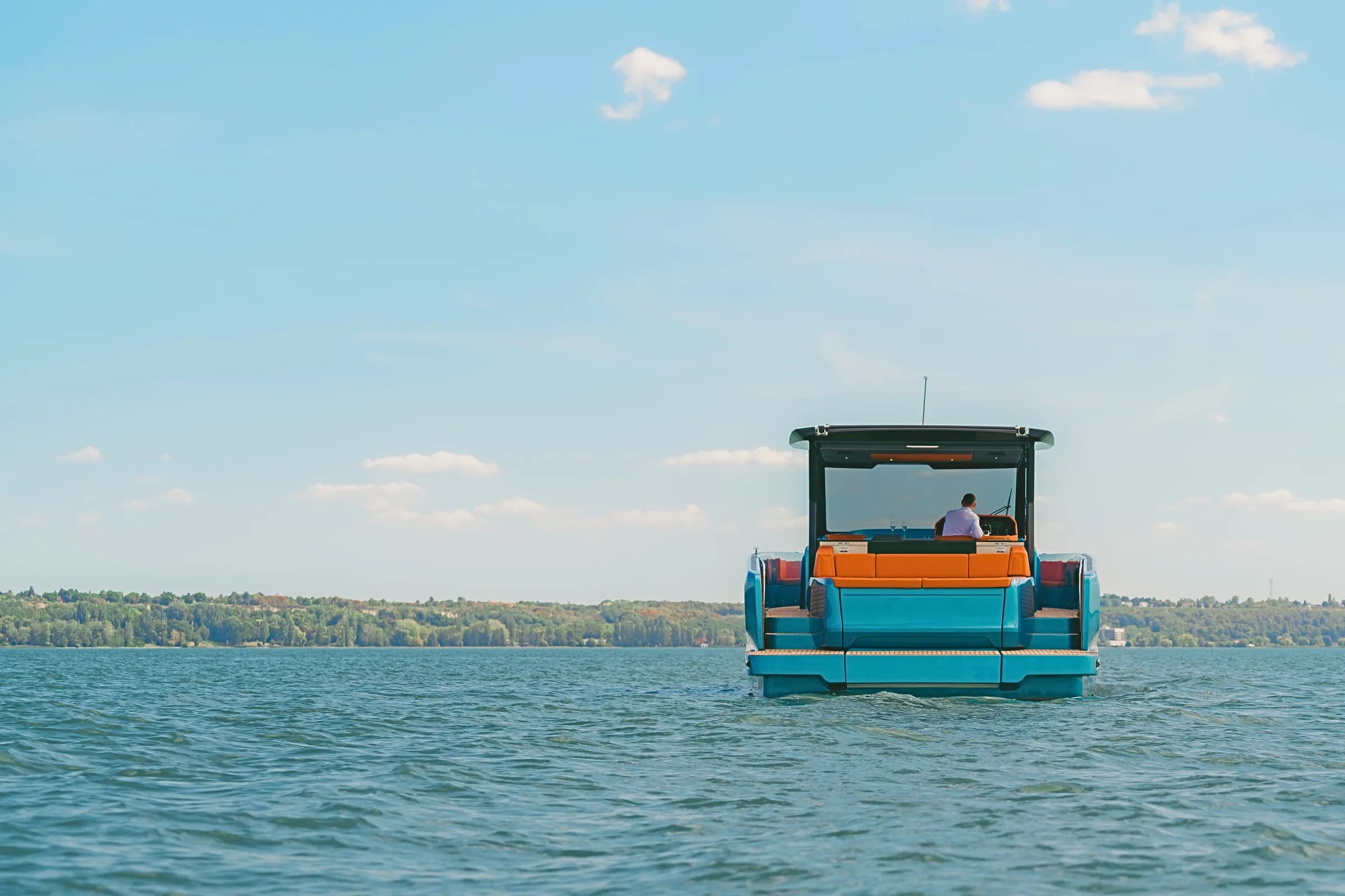 A turquoise-hulled Wia yacht seen from astern on a calm lake, its hardtop shading orange leather seating and a teak swim platform, one person at the helm, with a wooded shoreline on the horizon.