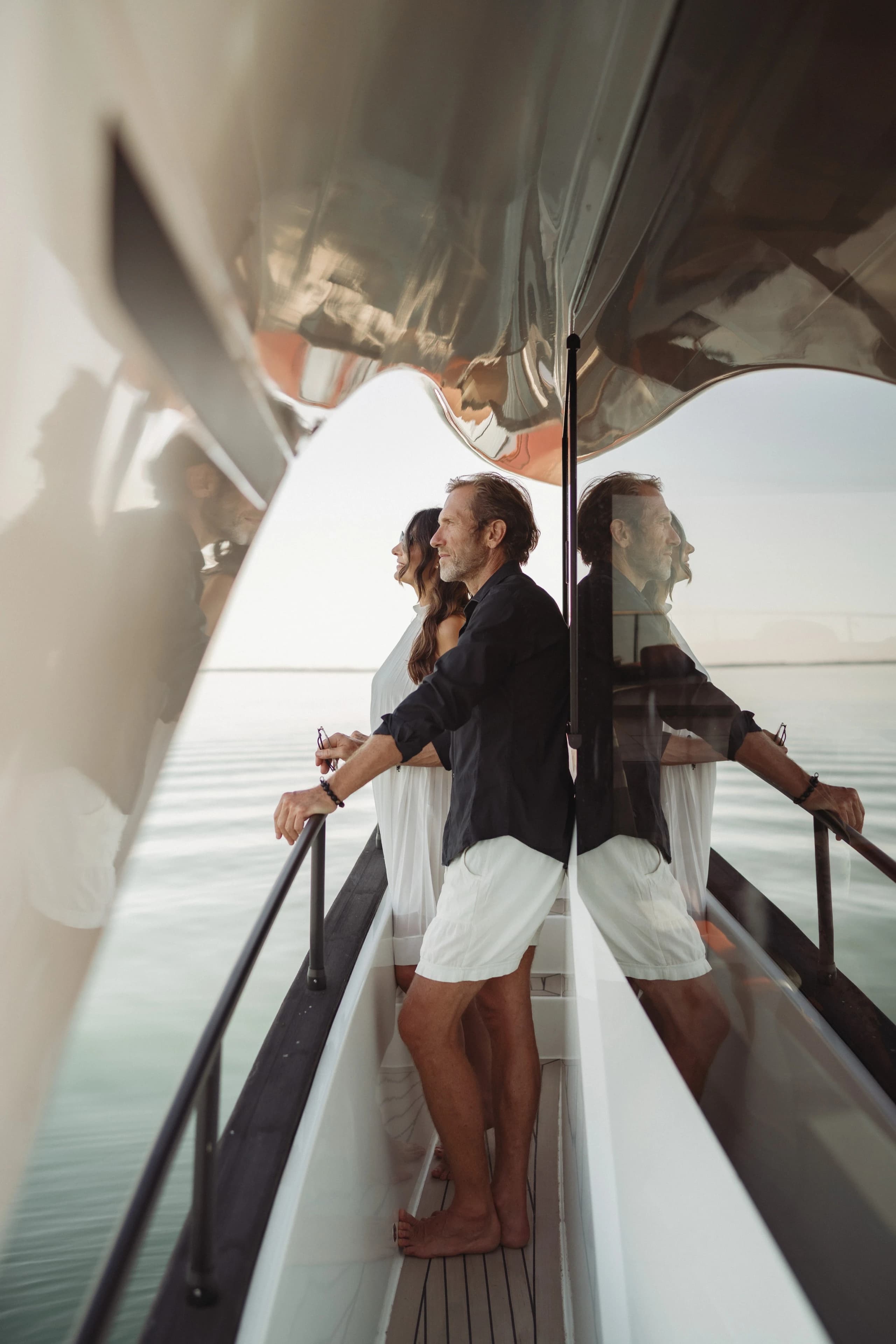 A couple standing at the railing on the side deck of a Wia yacht in hazy afternoon light.