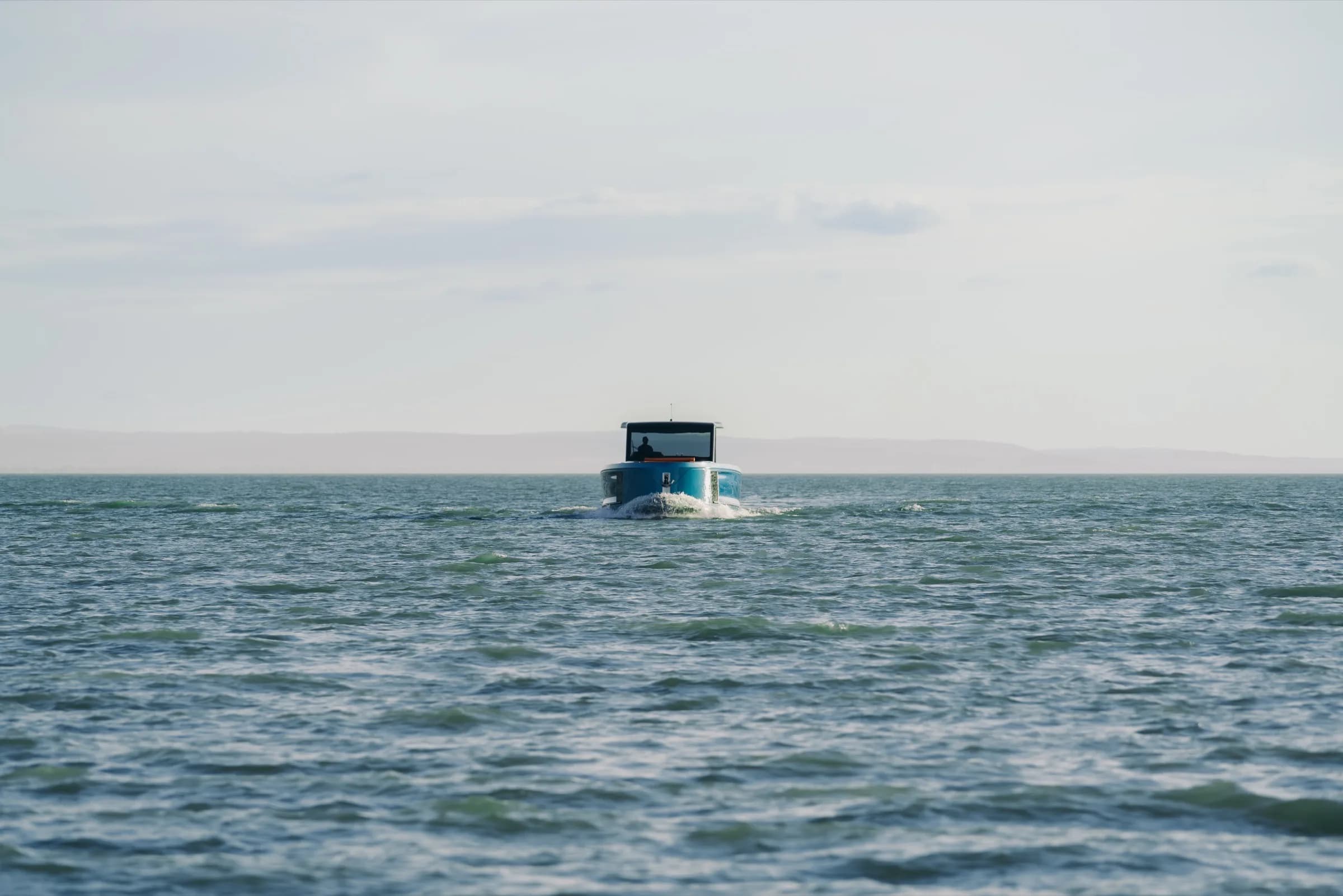 A blue-hulled Wia 520 Open cruising away across calm open water, seen from astern, a hazy shoreline on the horizon.
