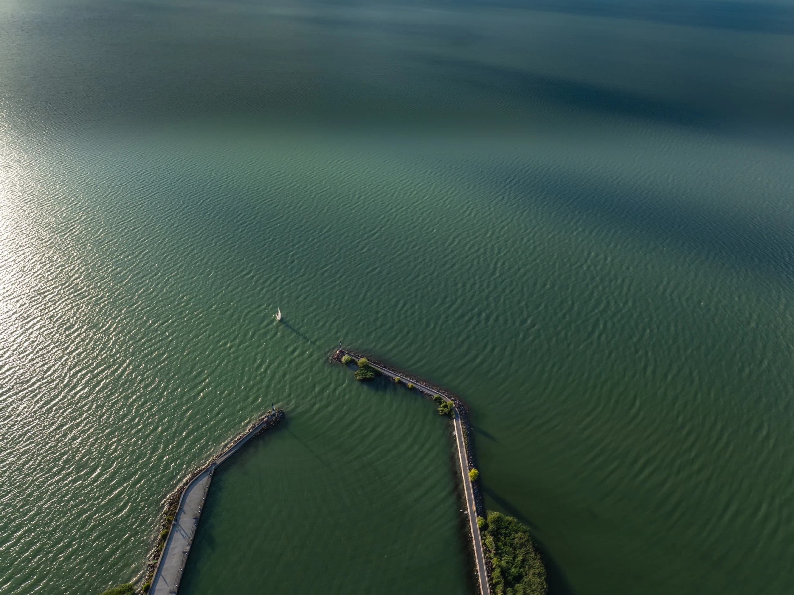 Aerial view of two stone breakwaters reaching into Lake Balaton, a lone sailboat between them.