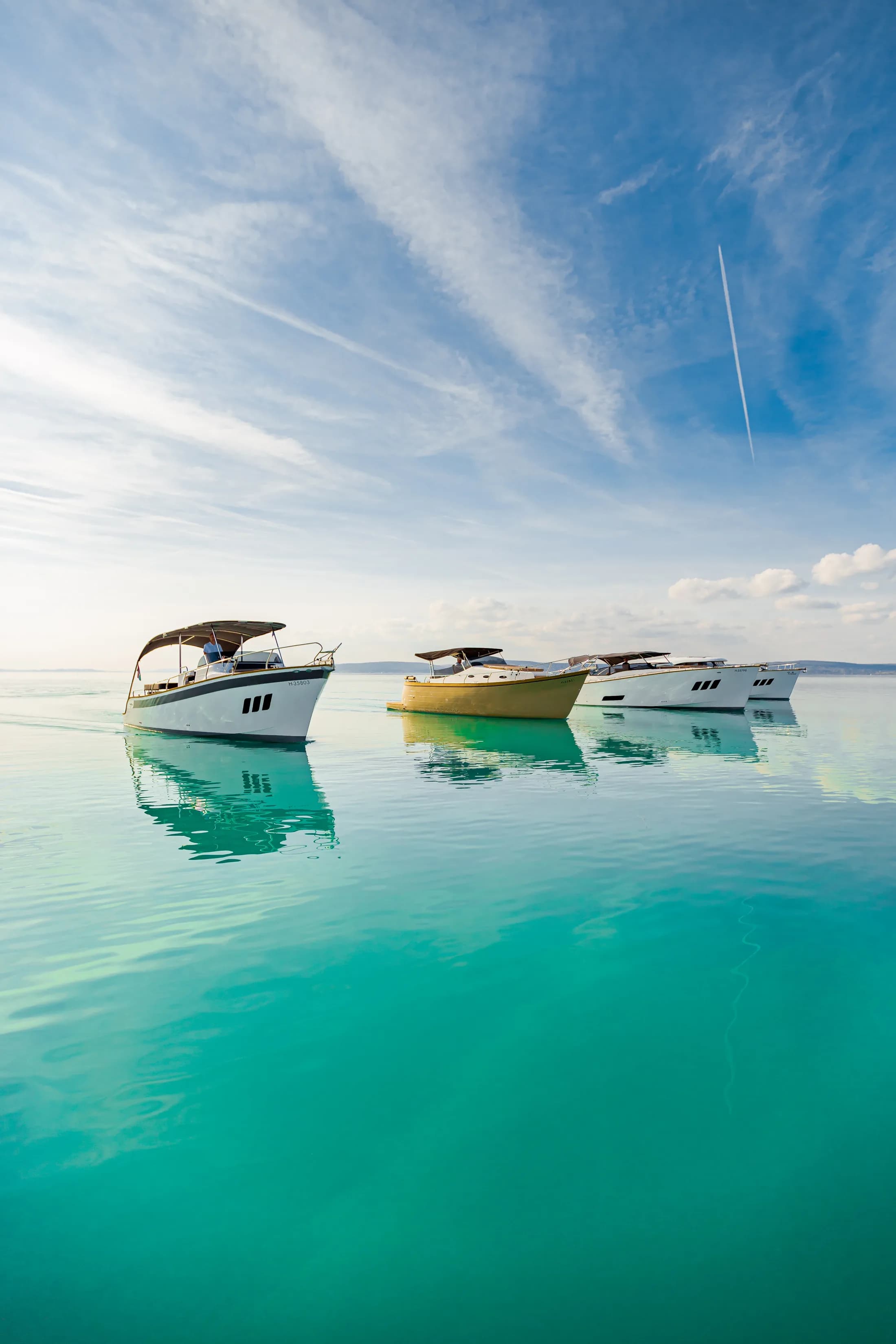 Four Wia yachts, one in yellow, floating in a row on calm turquoise water with clear reflections.