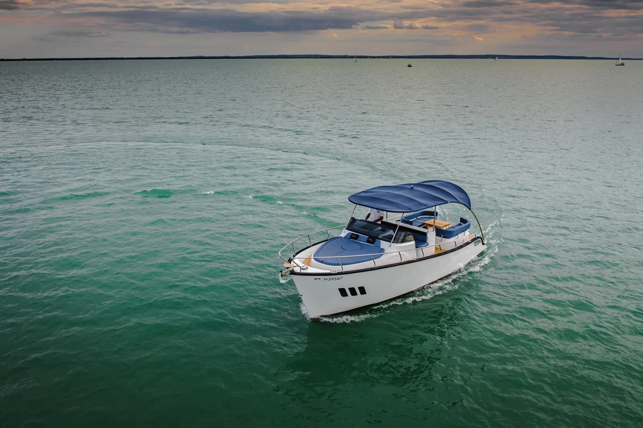 Aerial view of a white Wia with a blue bimini cruising alone across turquoise water at dusk.