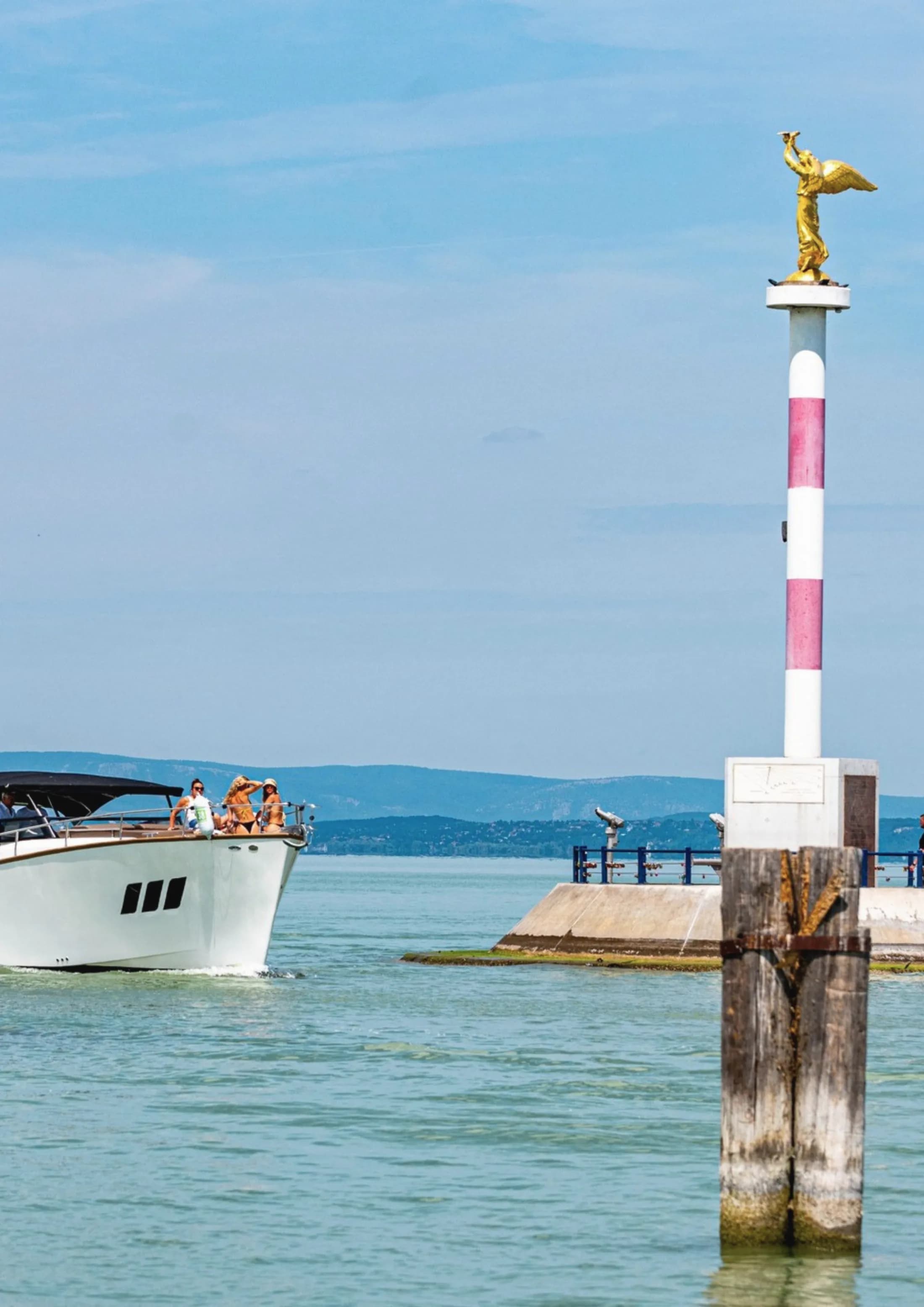 A white Wia with guests aboard cruising past a harbour entrance marked by a golden statue on a column.