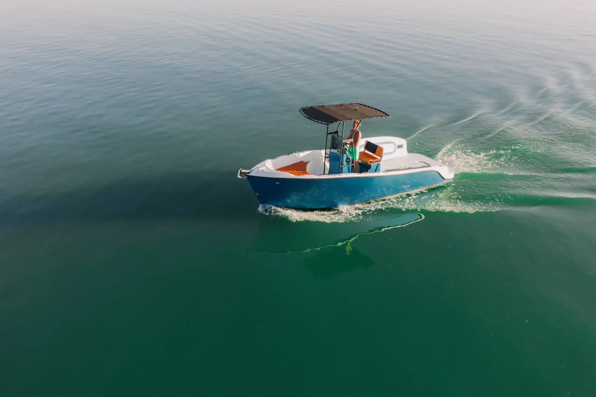 The blue-and-white-hulled Wia 230 CC centre-console boat with its black T-top under way on green water, a person at the console, seen from the air.