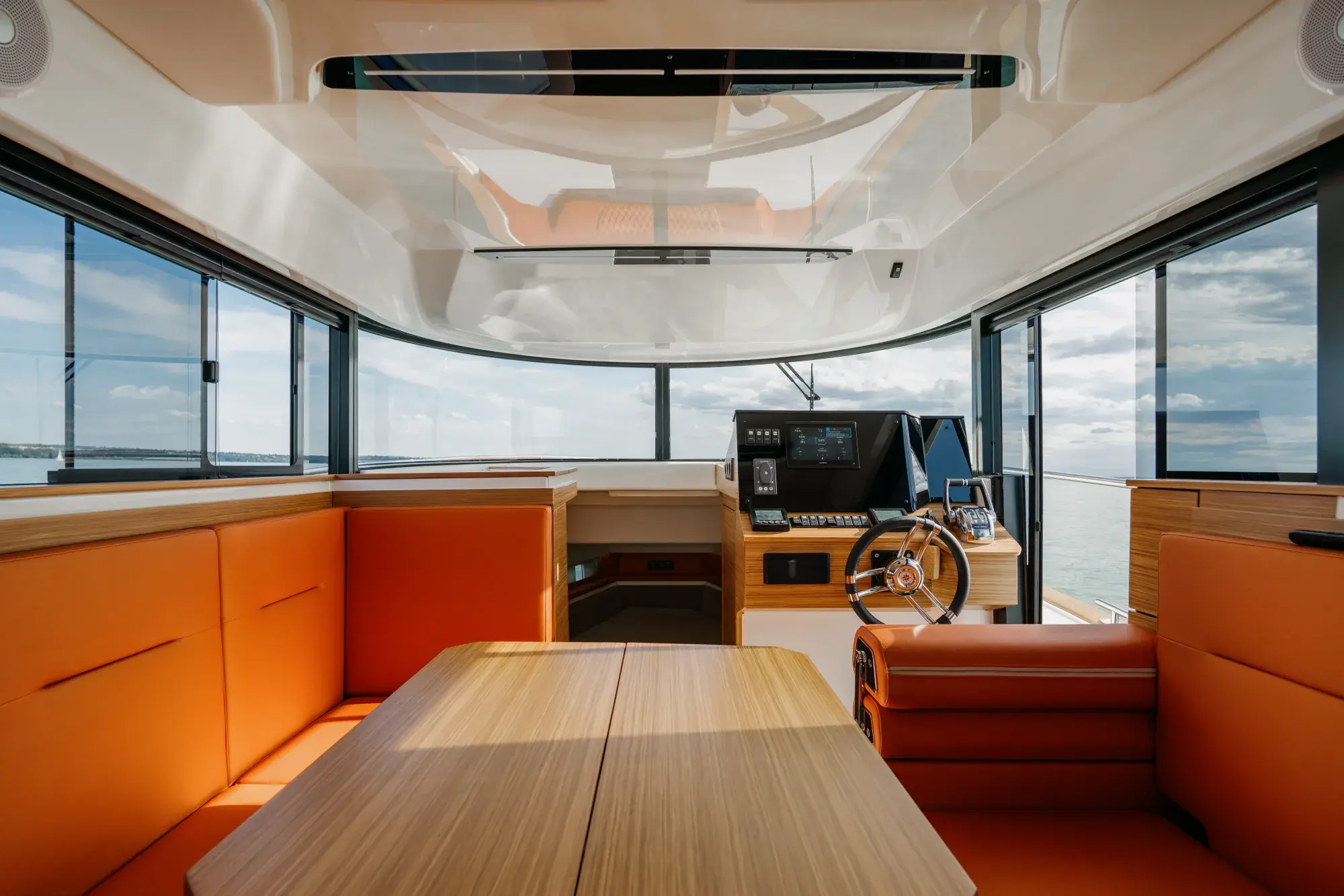 Das geschlossene Steuerhaus des Wia 350: leuchtend orangefarbene Ledersitze, eine Sitzgruppe aus heller Eiche, ein Steuerrad mit Chromspeichen und ein Touchscreen am Pult, umgeben von Panoramafenstern.
