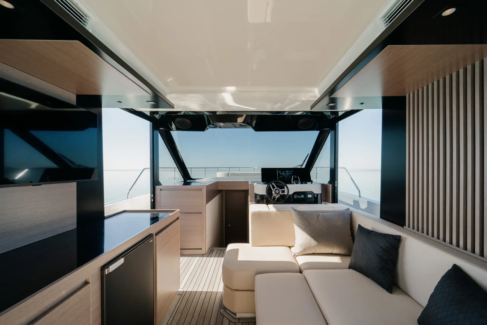 Der geschlossene Salon der Wia 520 HardTop: ein cremefarbenes L-Sofa, helle Eichenvertäfelung, eine Pantry mit dunkler Arbeitsplatte, ein Teakboden und der Steuerstand voraus, umgeben von Panoramafenstern.