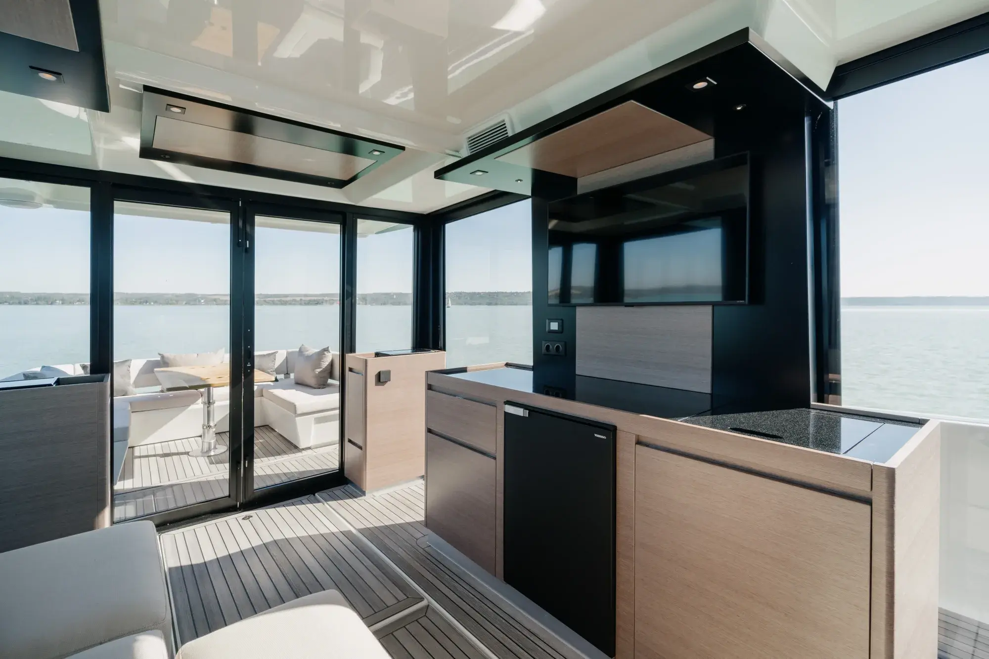 Der geschlossene Salon der Wia 520 HardTop: ein cremefarbenes L-Sofa, helle Eichenvertäfelung, eine Pantry mit dunkler Arbeitsplatte, ein Teakboden und der Steuerstand voraus, umgeben von Panoramafenstern.