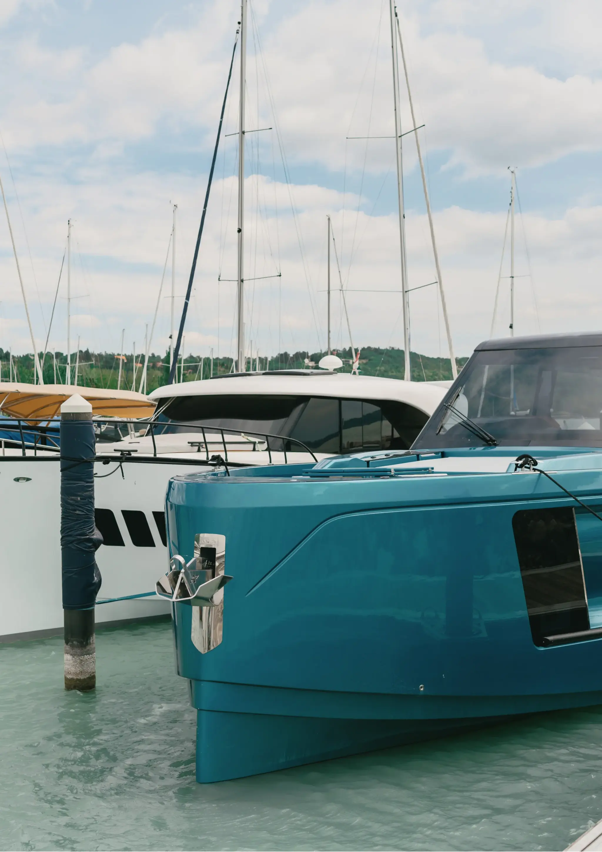 Der Bug einer petrolfarbenen Wia liegt in einer Marina neben einem gepolsterten Dalben, dahinter Segelbootmasten und bewaldete Hügel.