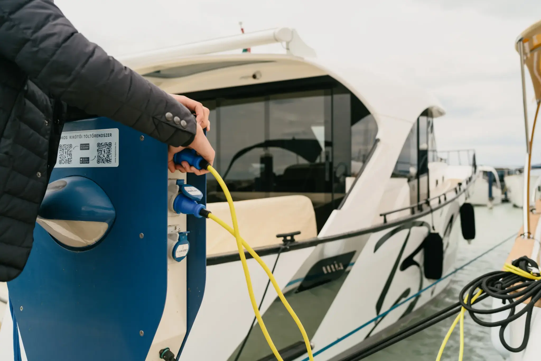 Eine Person steckt ein gelbes Landstromkabel in eine blaue Ladesäule der Marina neben einer vertäuten weißen Wia.