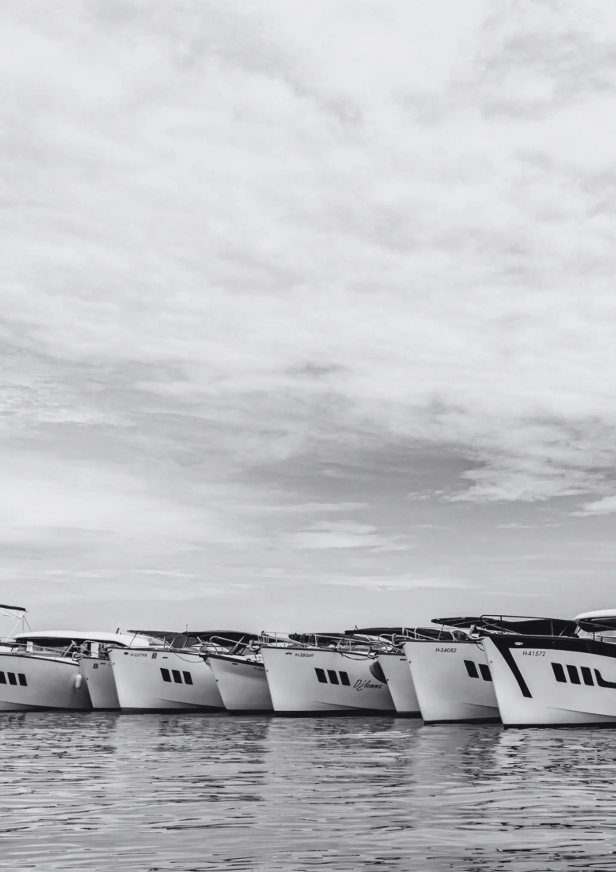 Black-and-white photo of a row of Wia yachts moored bow to bow on the water under a cloudy sky.