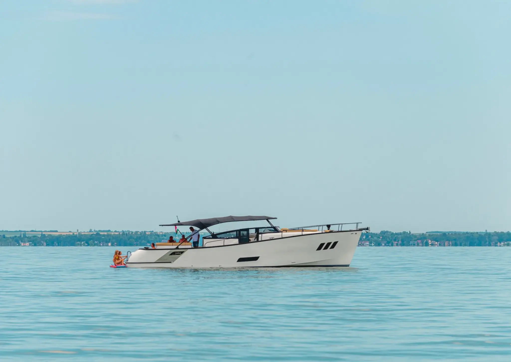 A white Wia yacht cruising in profile on a calm blue lake, guests under a dark bimini top and two people on a paddleboard at the stern.
