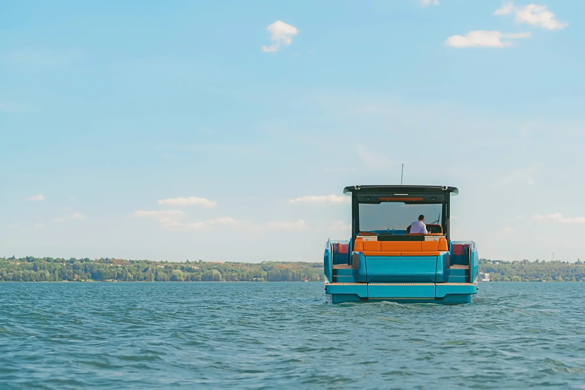 A turquoise-hulled Wia yacht seen from astern on a calm lake, its hardtop shading orange leather seating and a teak swim platform, one person at the helm, with a wooded shoreline on the horizon.