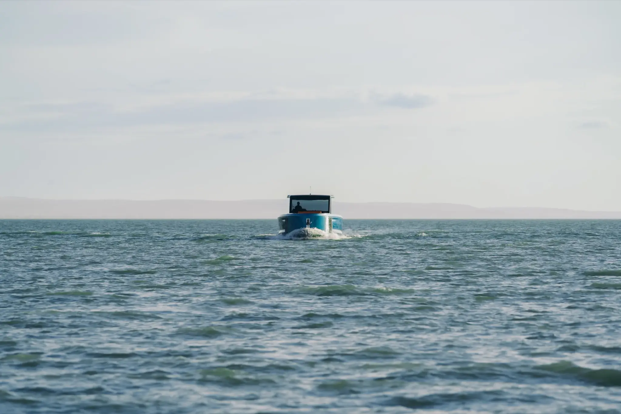 A blue-hulled Wia 520 Open cruising away across calm open water, seen from astern, a hazy shoreline on the horizon.