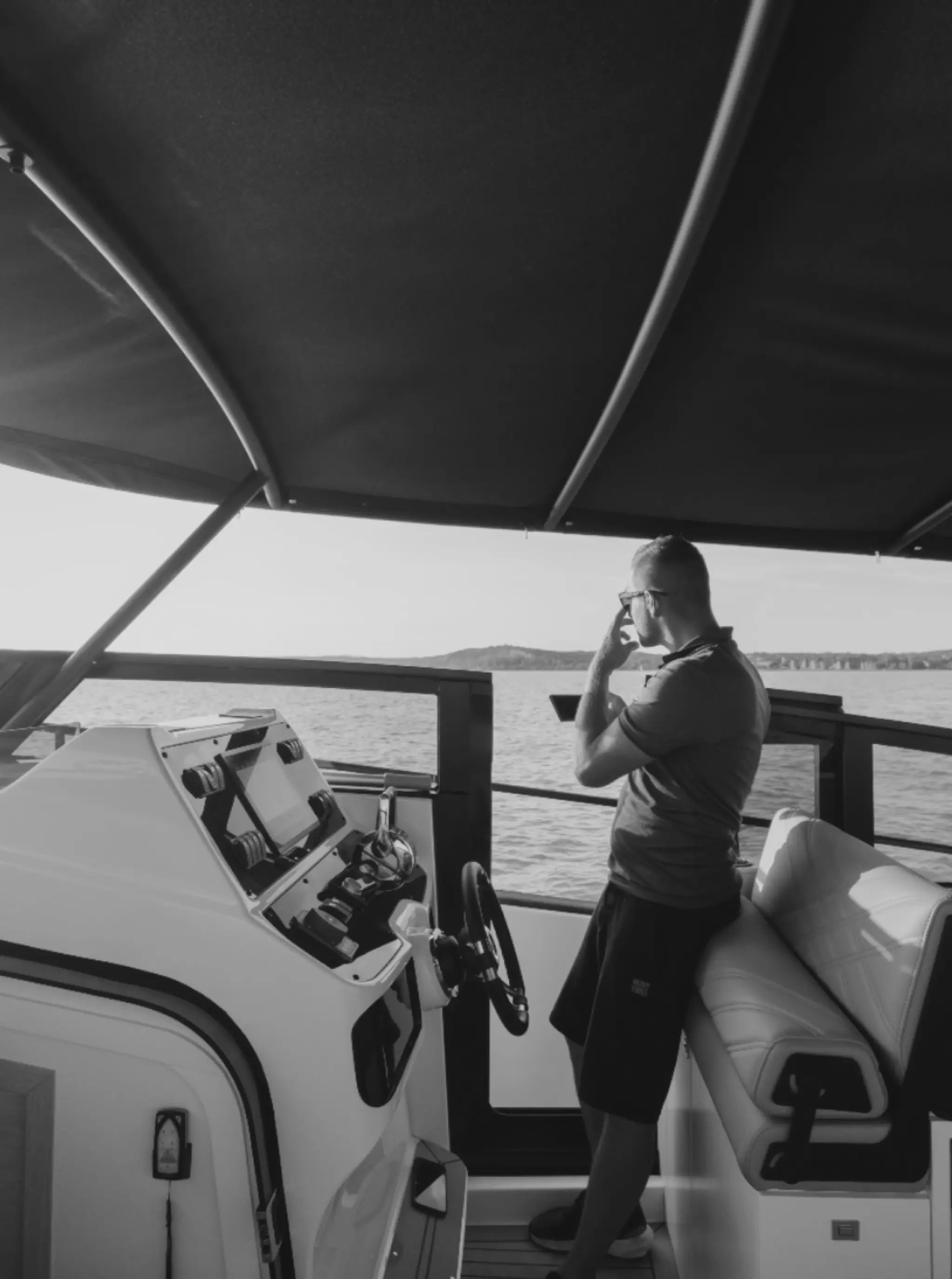 Black-and-white photo of a man at the helm of a Wia yacht under the bimini top, scanning the water ahead.